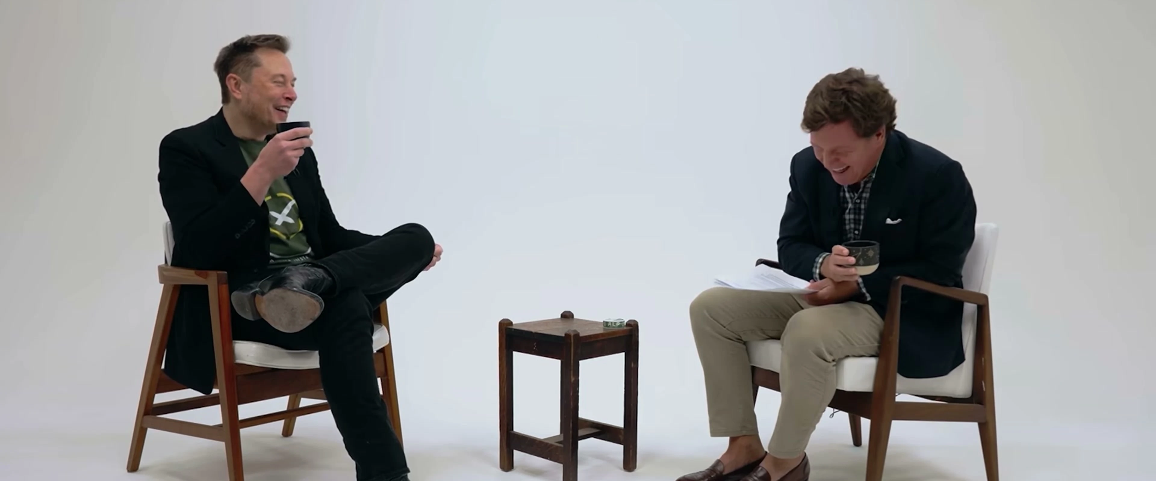 Elon Musk laughs while sitting across from another man in a bright scene from the documentary trailer.