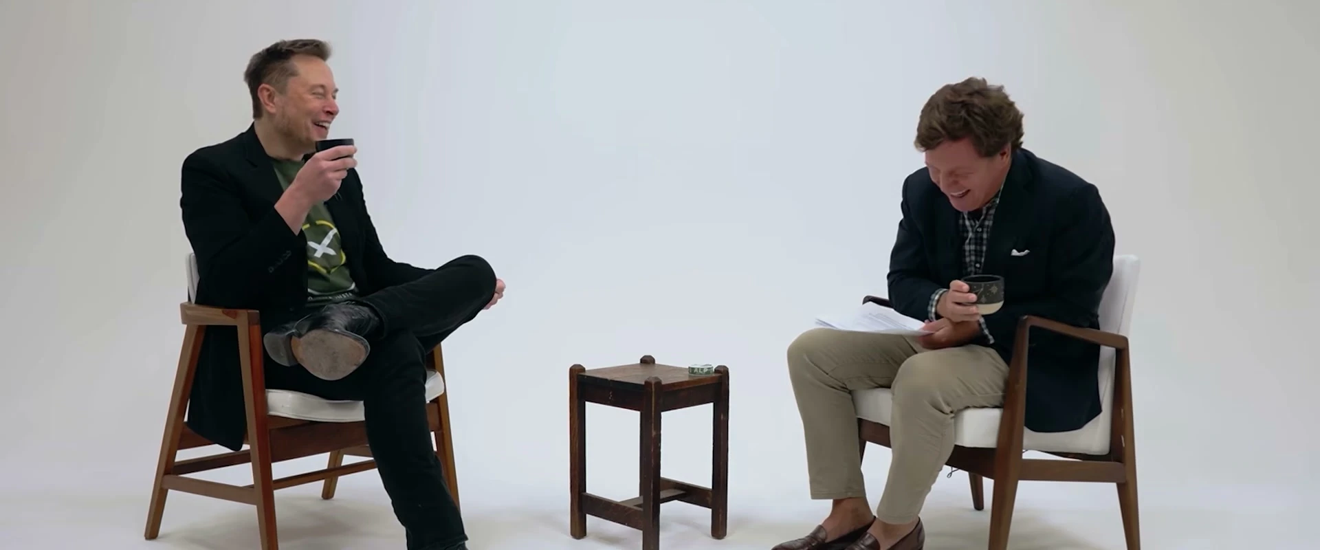 Elon Musk laughs while sitting across from another man in a bright scene from the documentary trailer.
