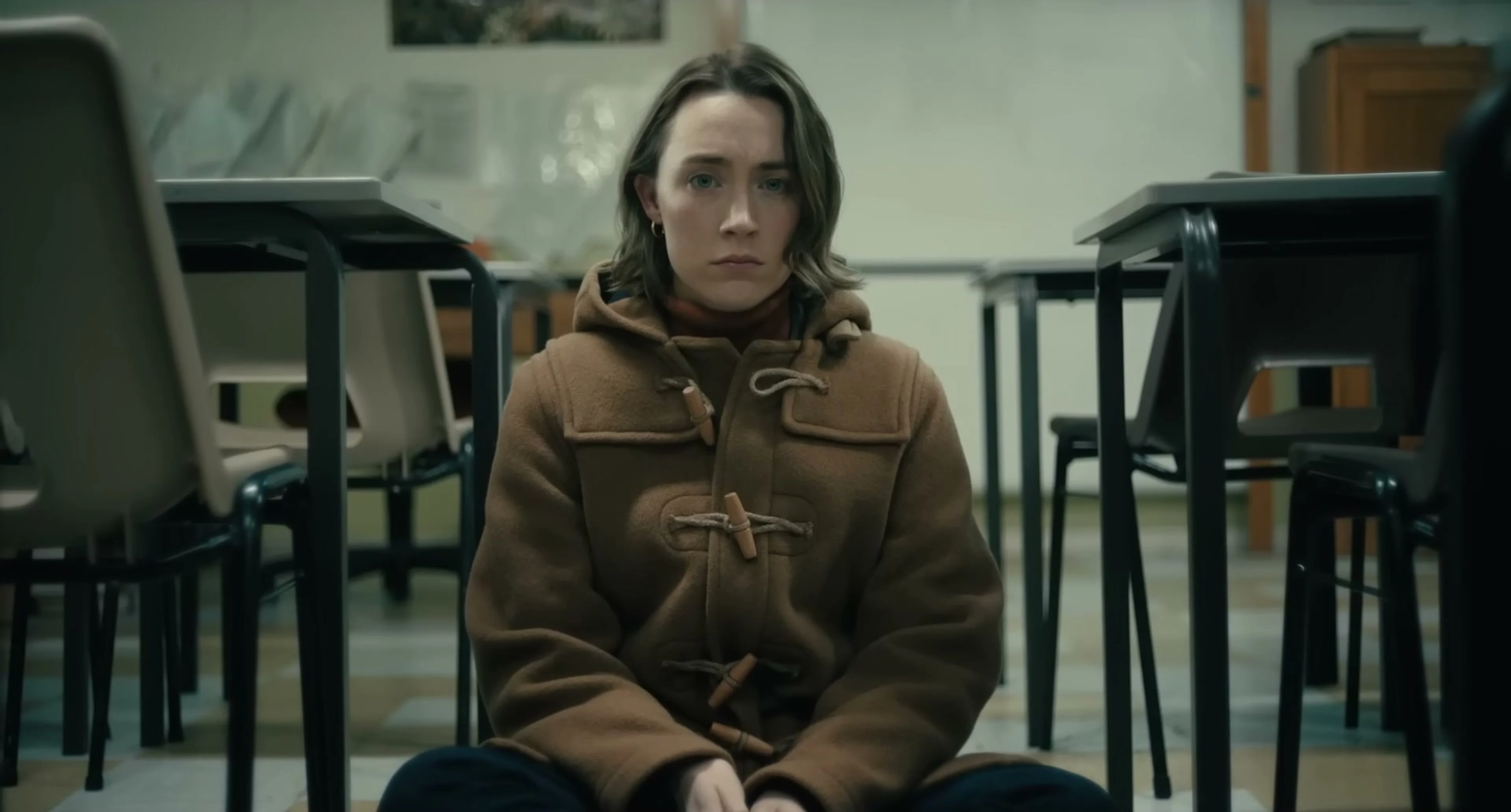 Maria sits alone in a classroom wearing a brown coat.