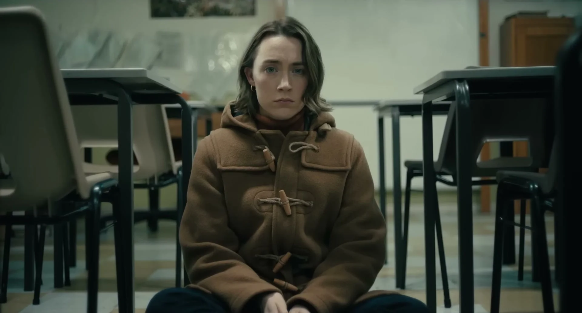 Maria sits alone in a classroom wearing a brown coat.
