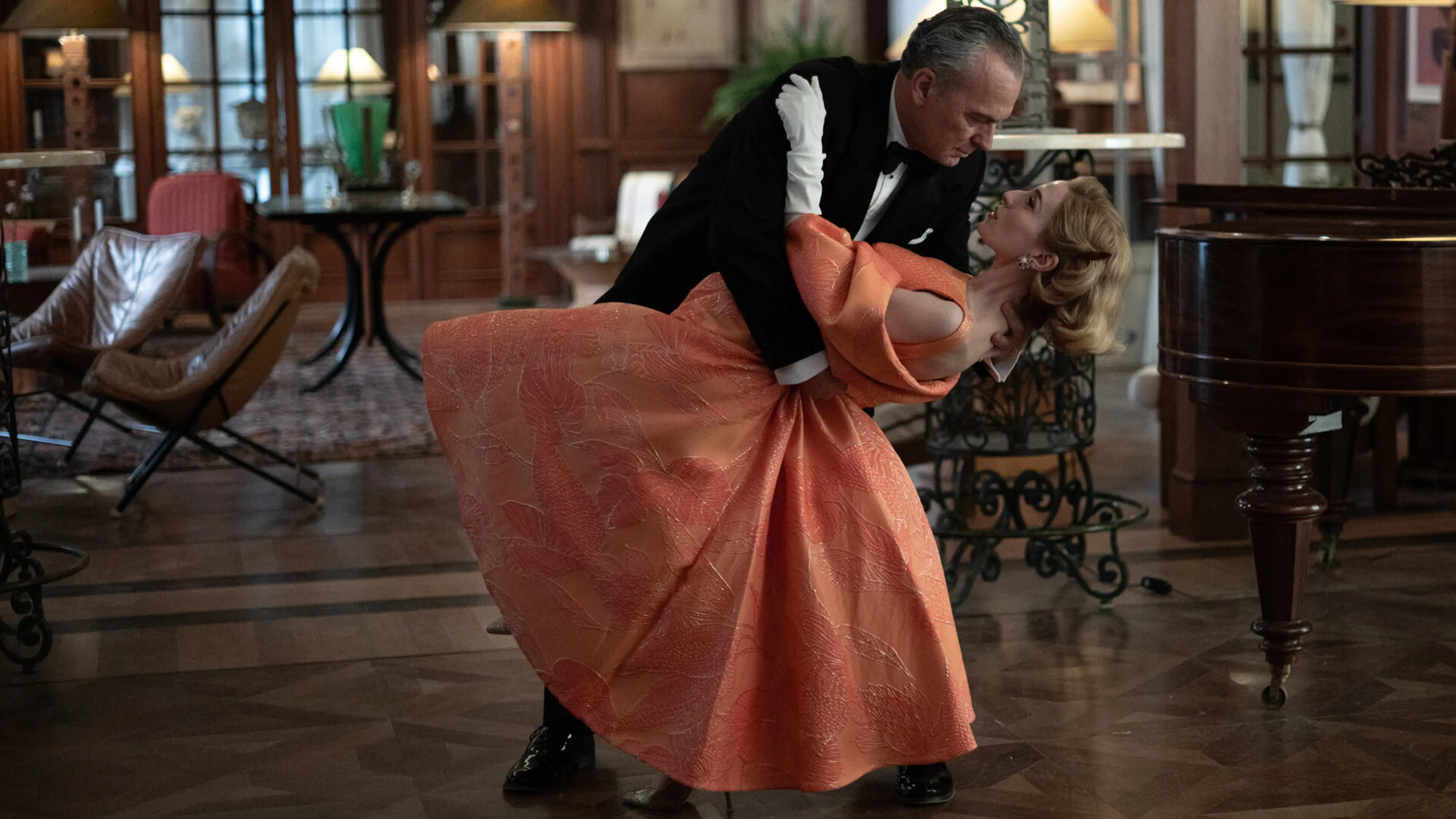 Basil dances with a woman in a peach-colored dress beside a piano.