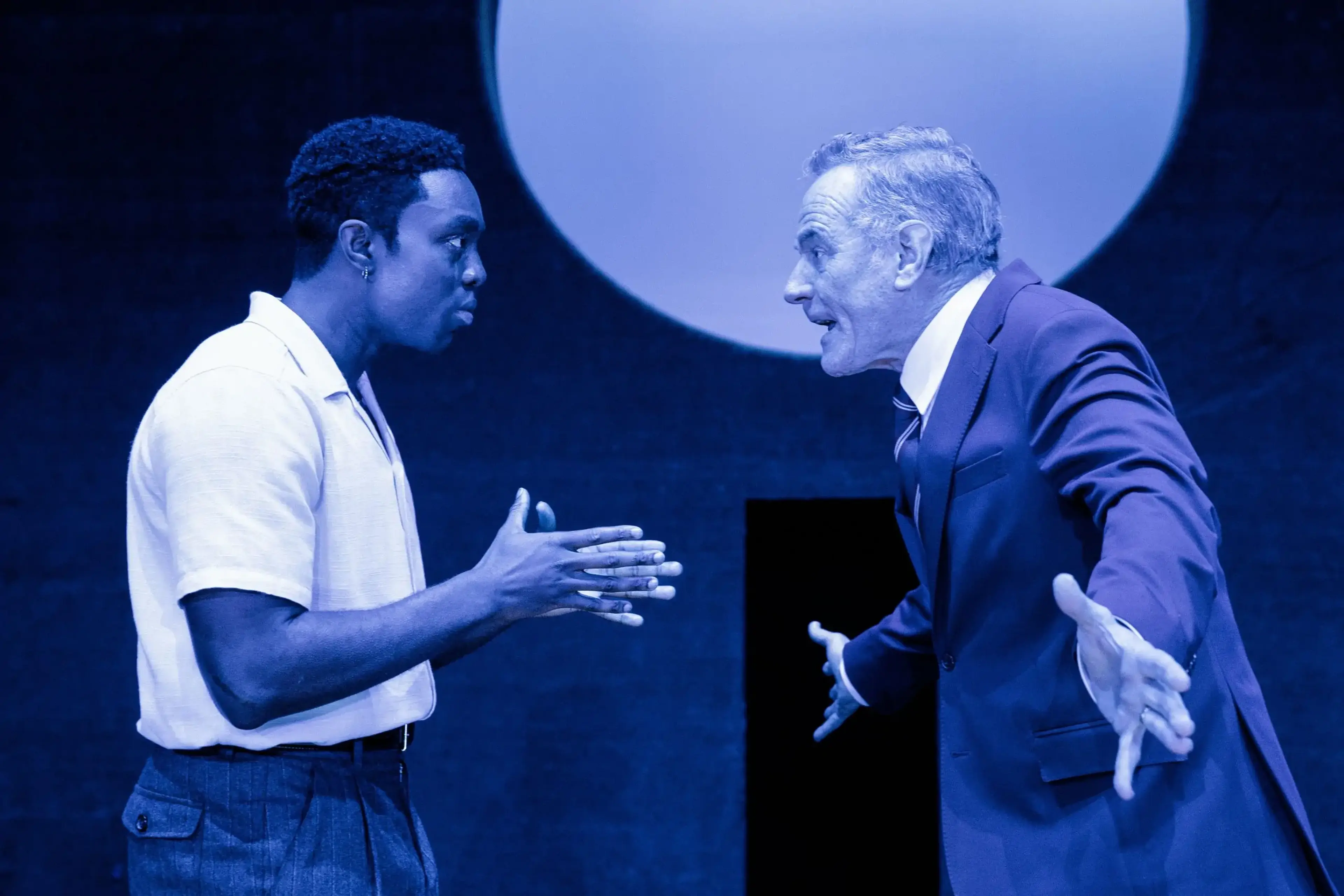 Paapa Essiedu and Bryan Cranston face each other under blue stage lighting in All My Sons.