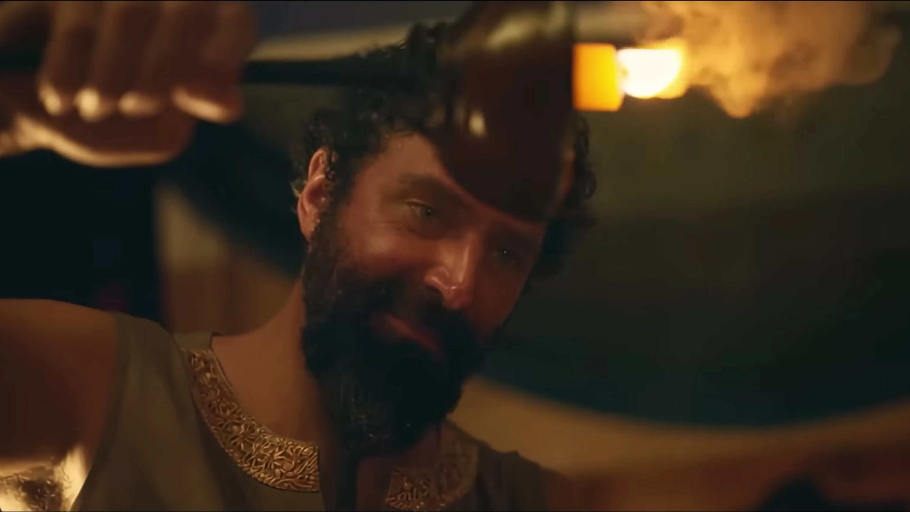 Elijah Alexander as King Nebuchadnezzar watches someone closely in a torchlit room.