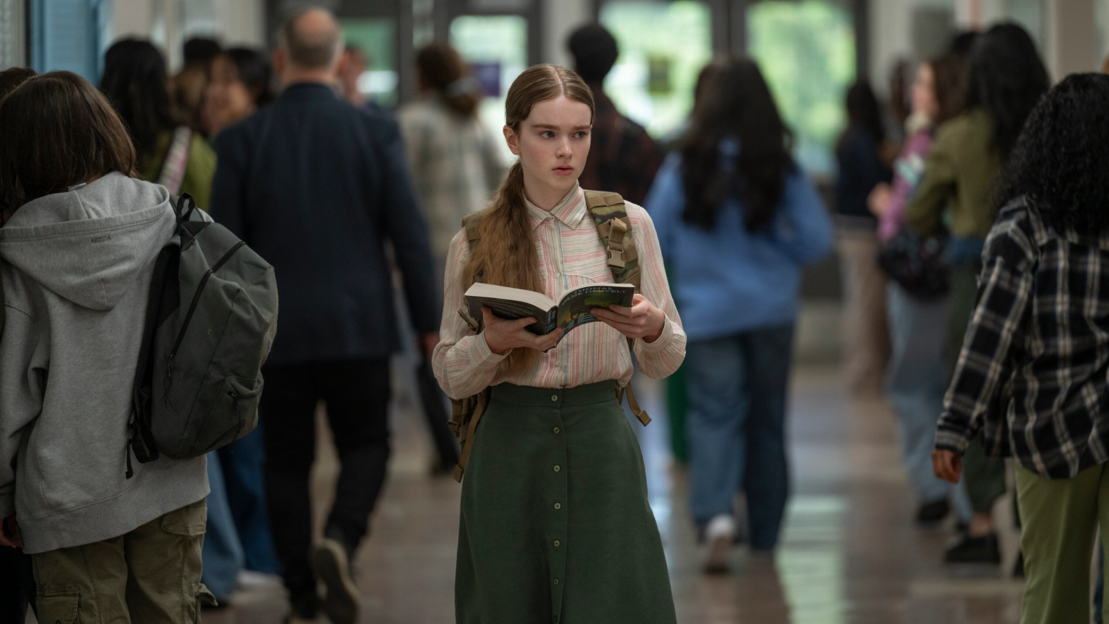 Carrie stands in a school corridor holding a book and looking to her right.