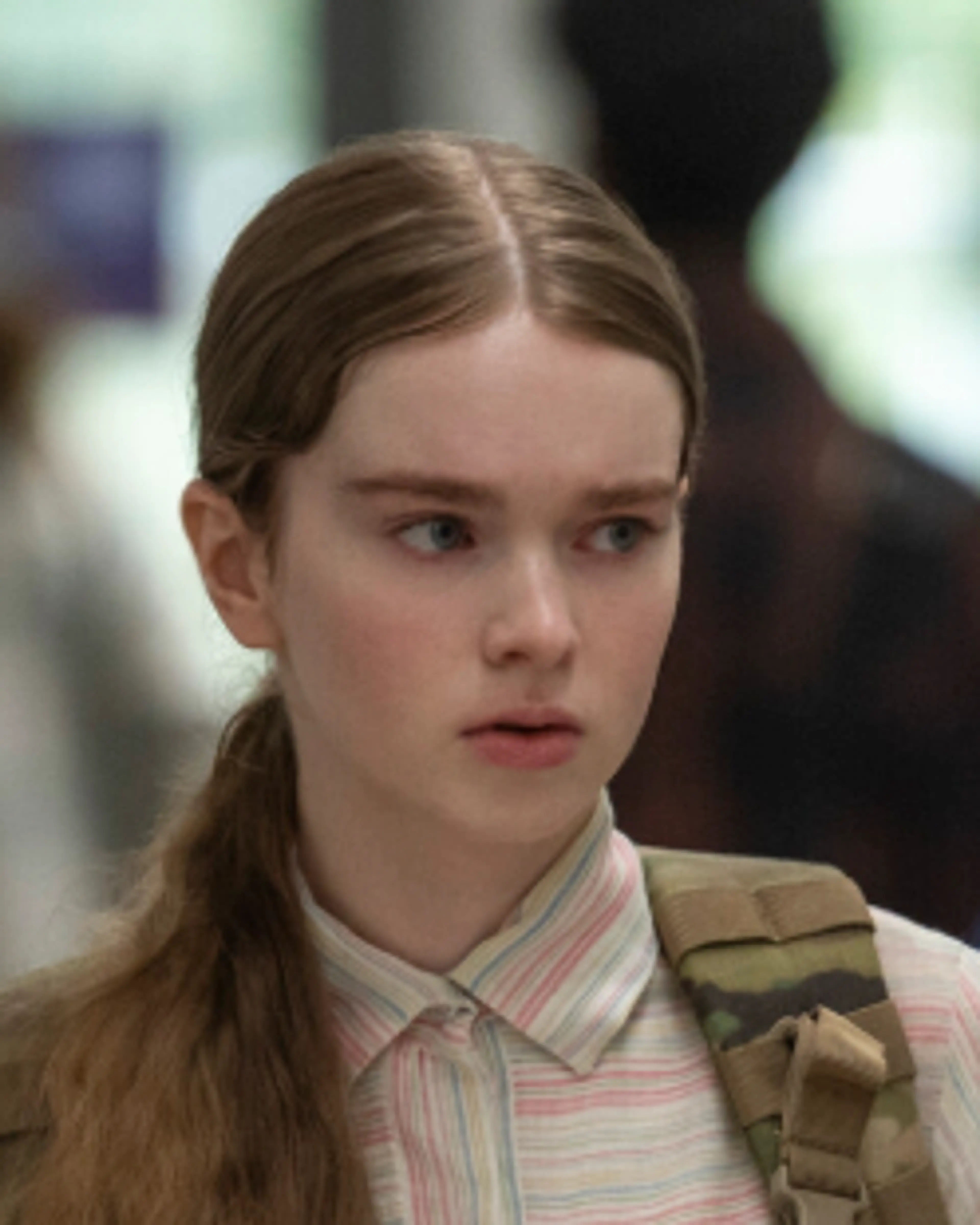 Carrie stands in a school corridor holding a book and looking to her right.