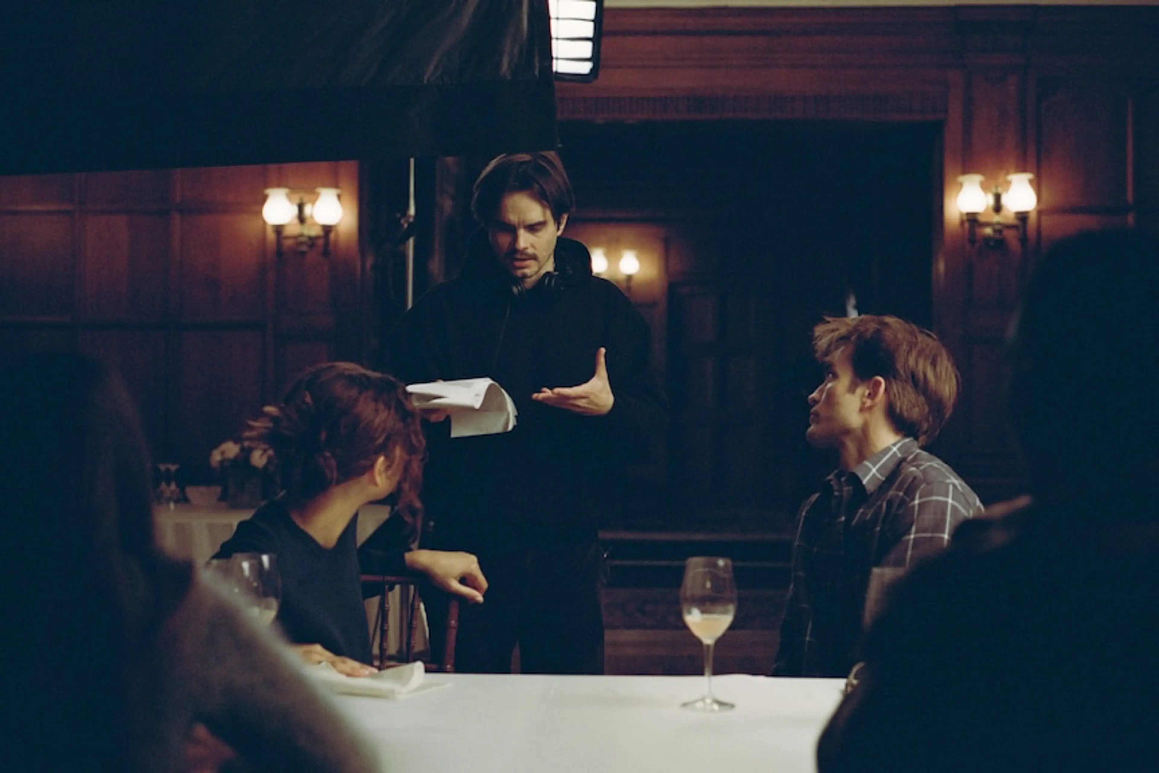Director Kristoffer Borgli stands holding pages between Zendaya and Robert Pattinson at a dining table.