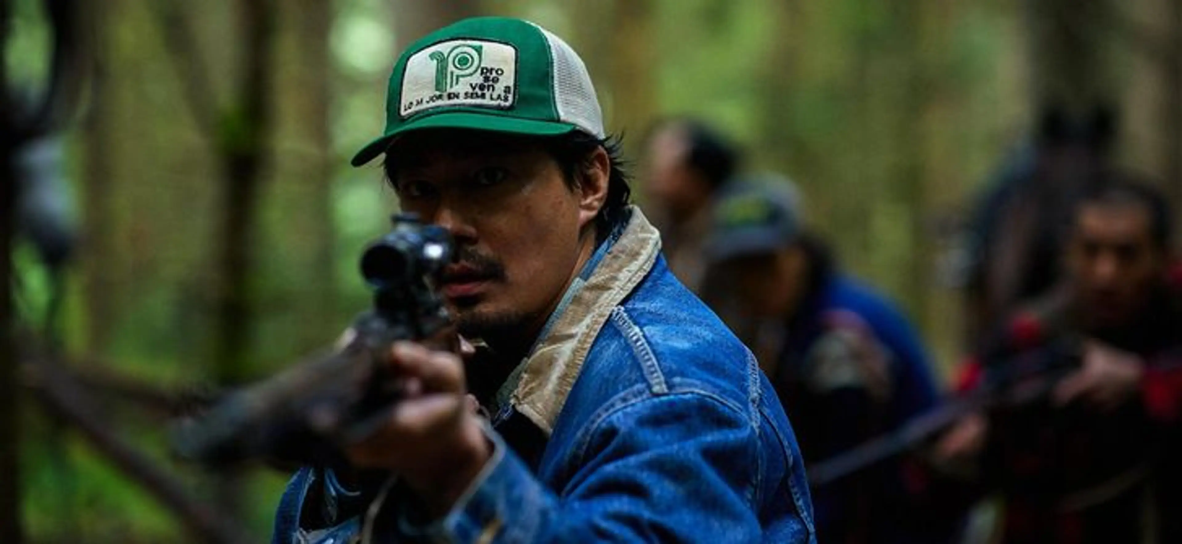 Sung-ki aims a rifle in a forest, with other hunters behind him.
