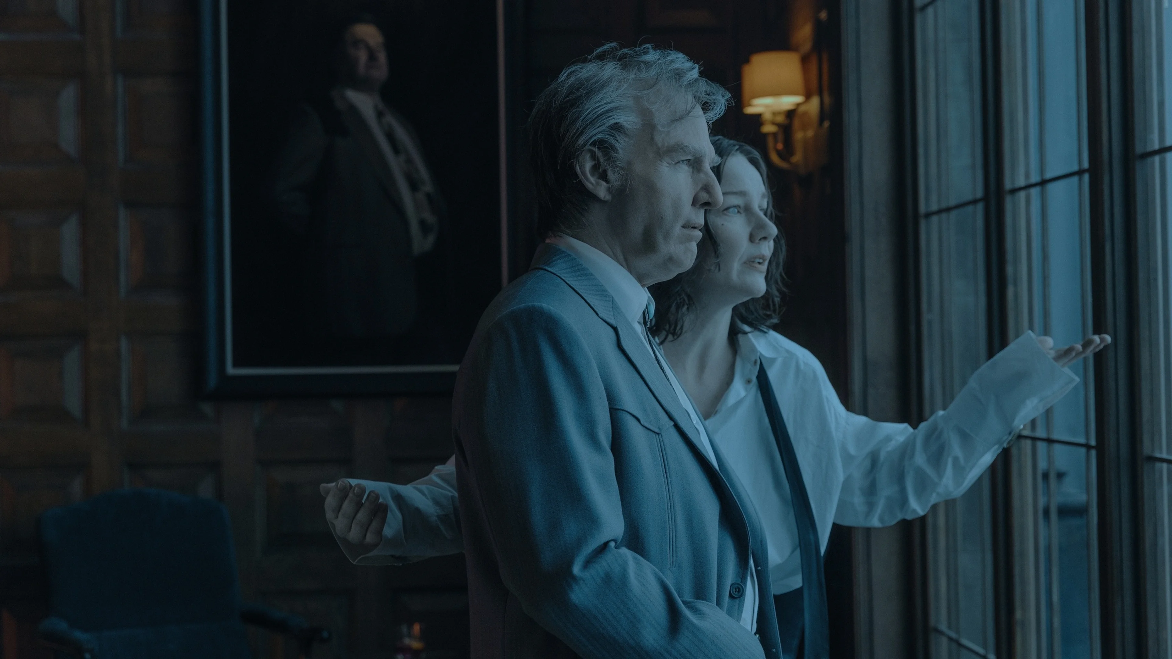 Tom Cruise stands beside a large window in a dim blue-lit room in Digger.