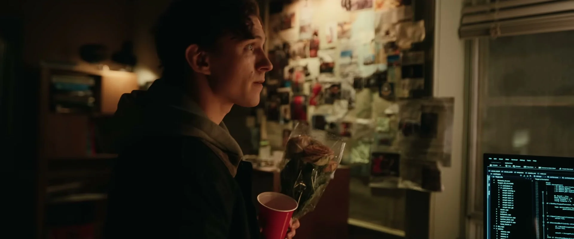 Tom Holland as Peter Parker holds flowers and a red cup beside a wall of Spider-Man pictures and a computer.