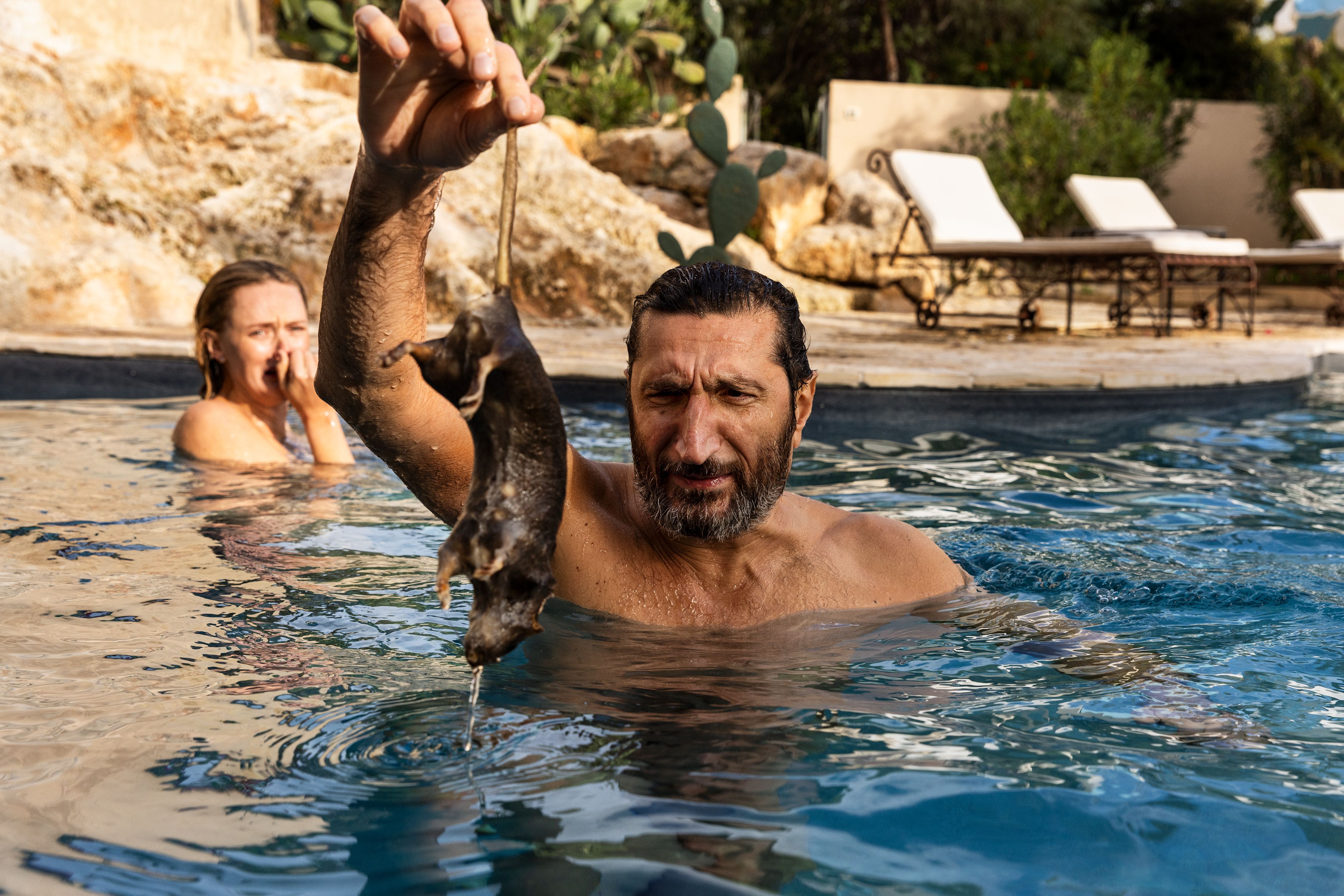 Fares Fares as Carl holds up a fish while standing in a swimming pool in A Couple of Lies.