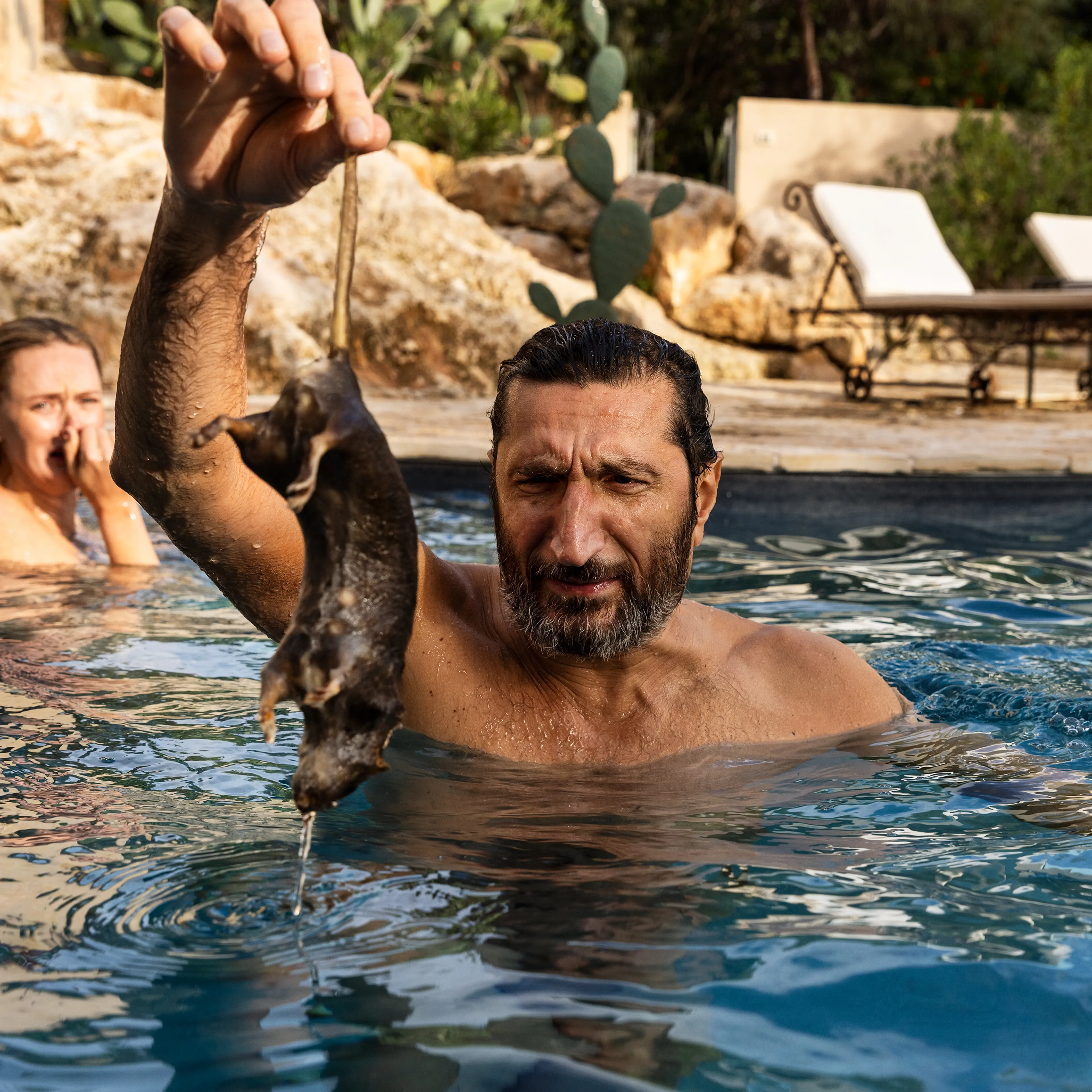 Fares Fares as Carl holds up a fish while standing in a swimming pool in A Couple of Lies.