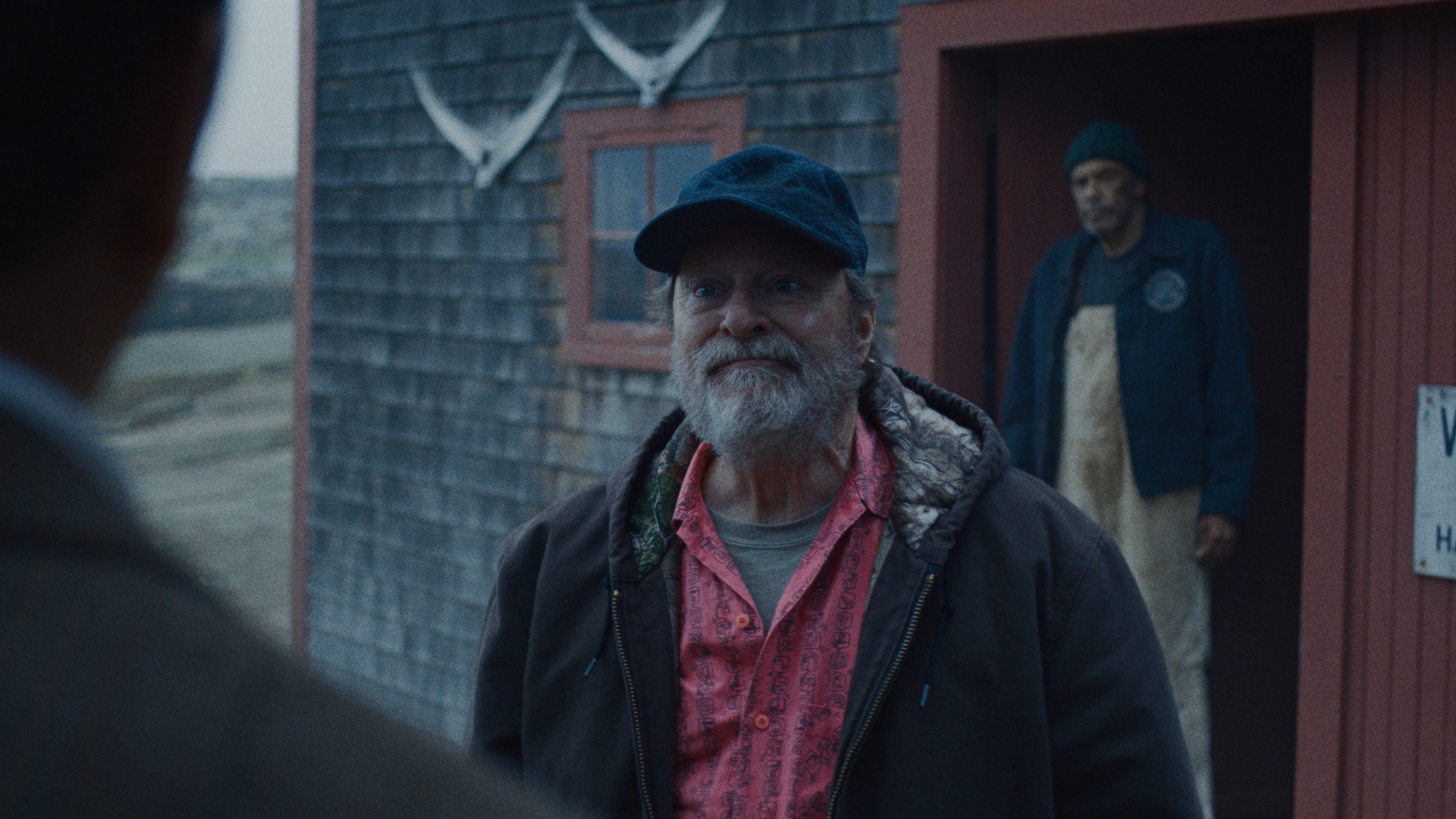 Stephen Root as Wyck, wearing a cap and red shirt, speaks outside a building.