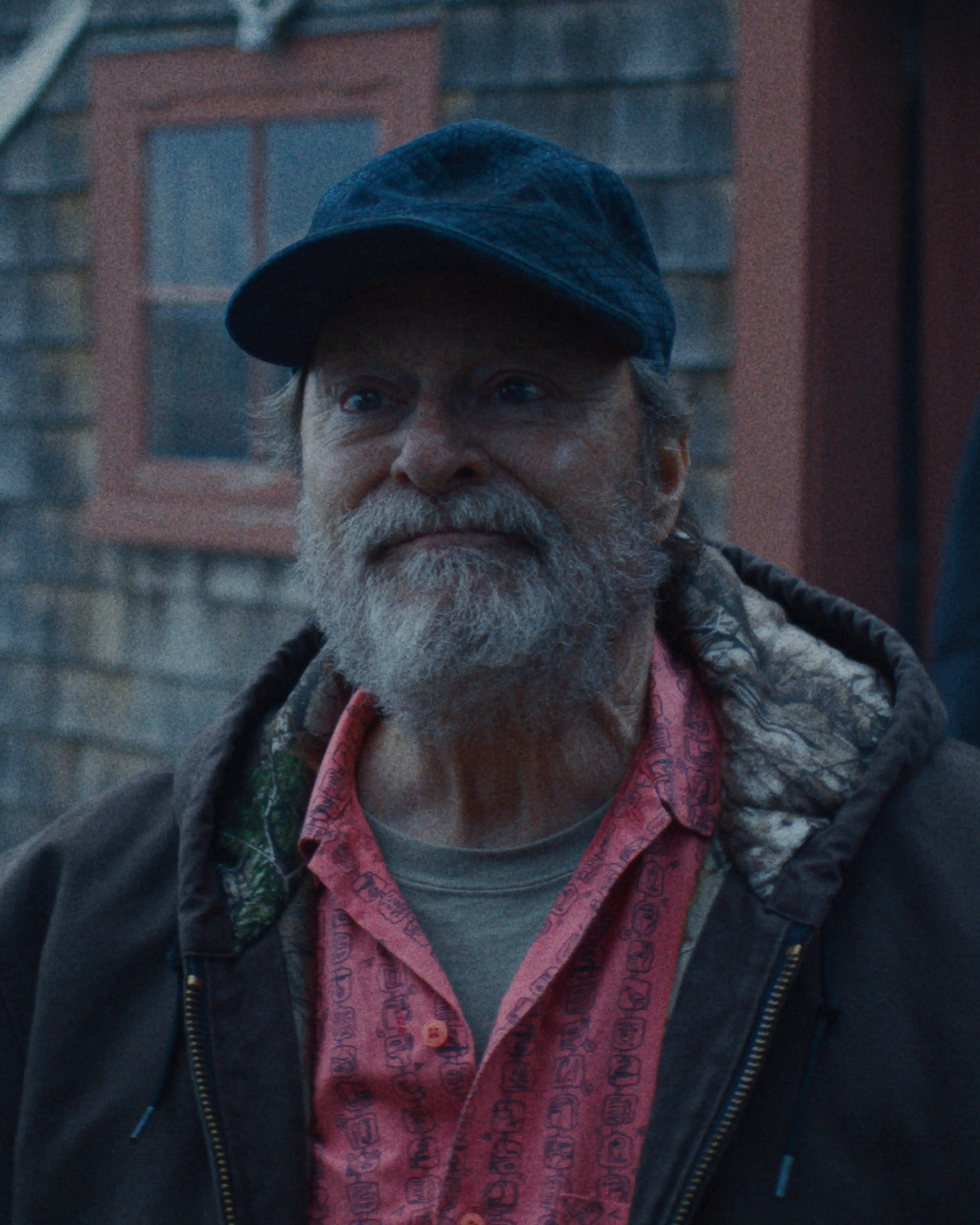 Stephen Root as Wyck, wearing a cap and red shirt, speaks outside a building.
