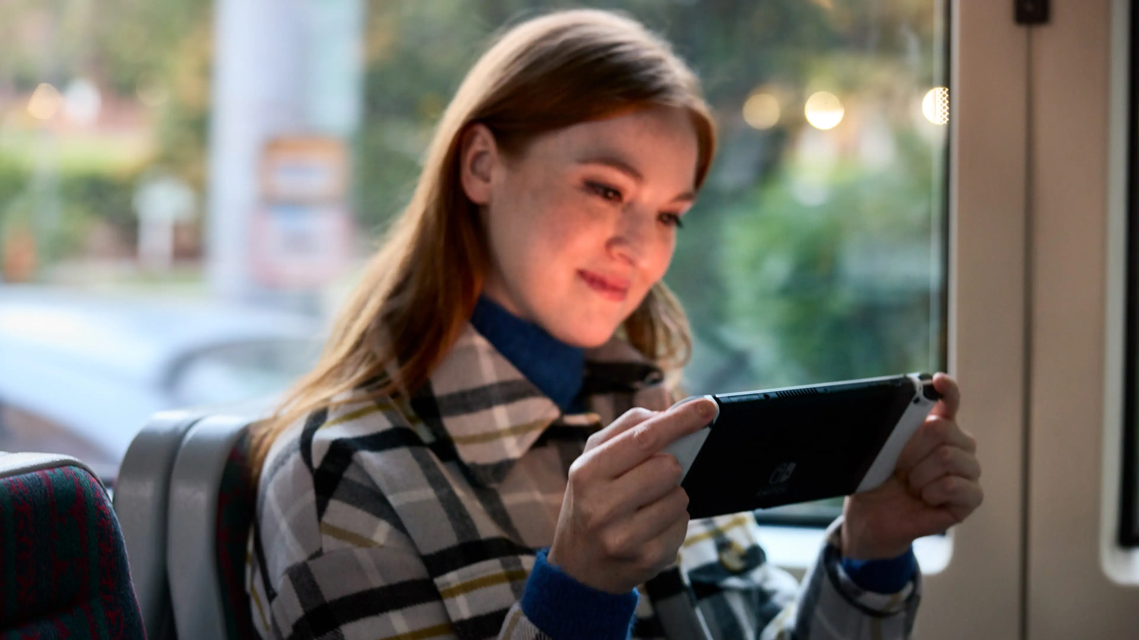 A person uses a white Nintendo Switch OLED Model in handheld mode on a train.