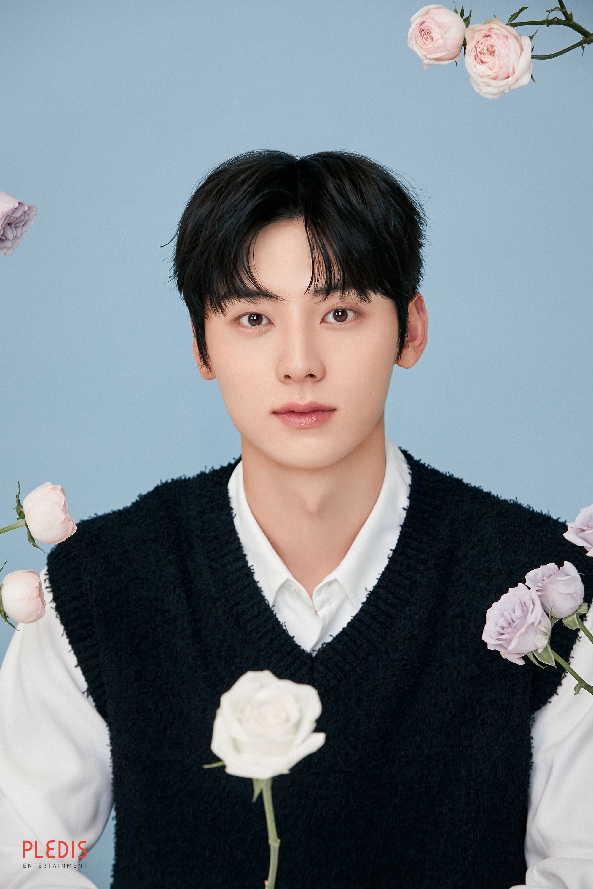 Hwang Minhyun facing the camera with pale roses, wearing a black knit vest over a white shirt against a light blue background.