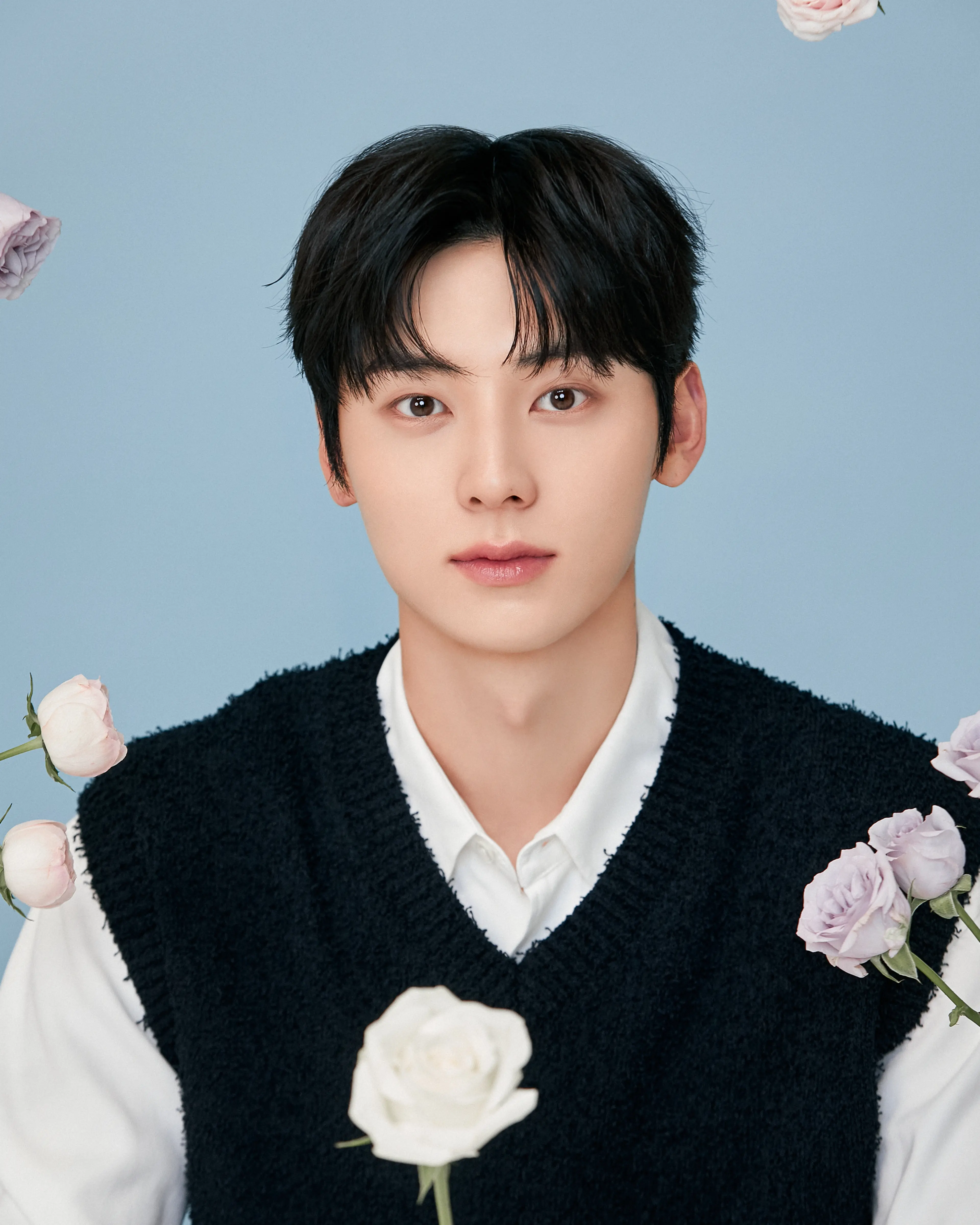 Hwang Minhyun facing the camera with pale roses, wearing a black knit vest over a white shirt against a light blue background.