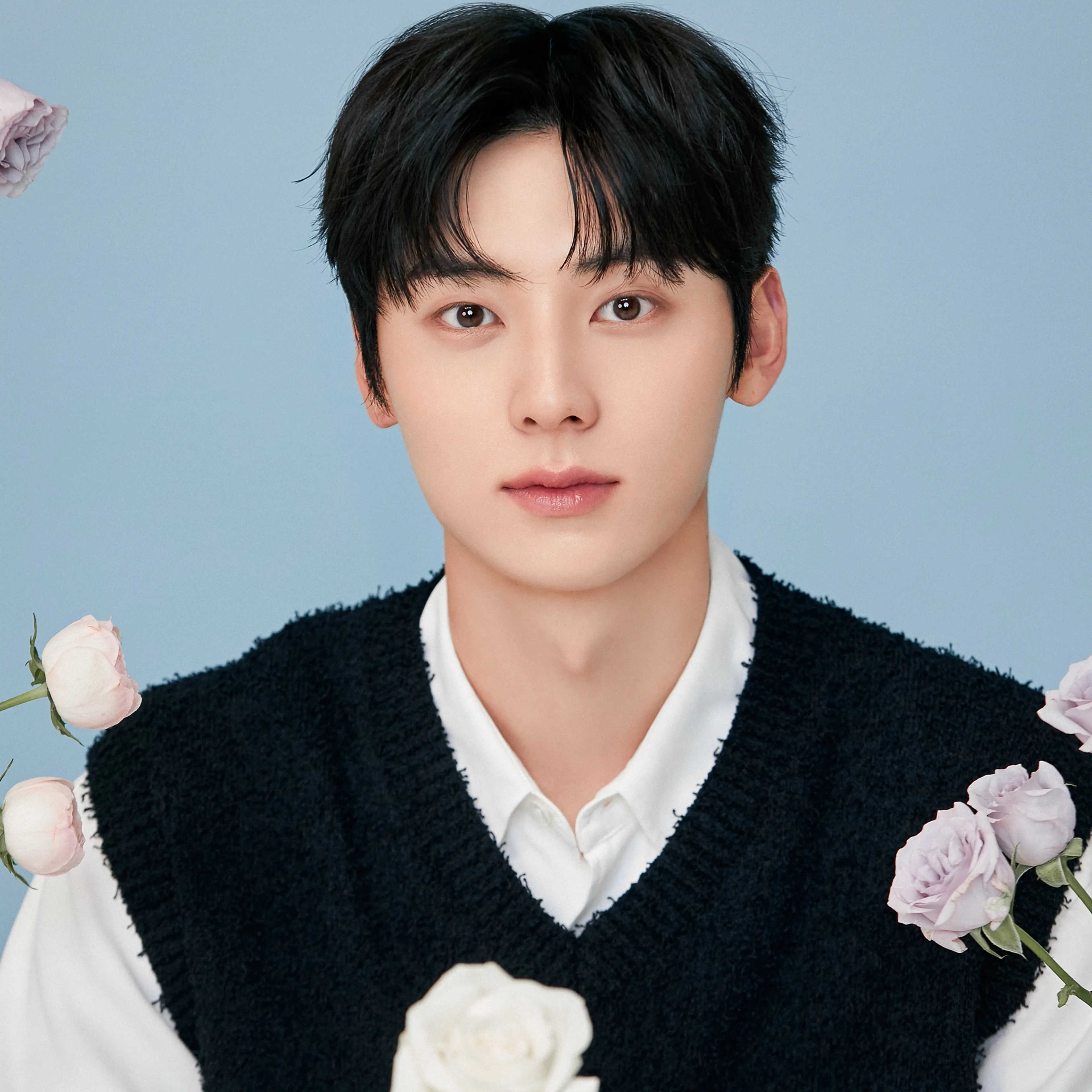 Hwang Minhyun facing the camera with pale roses, wearing a black knit vest over a white shirt against a light blue background.