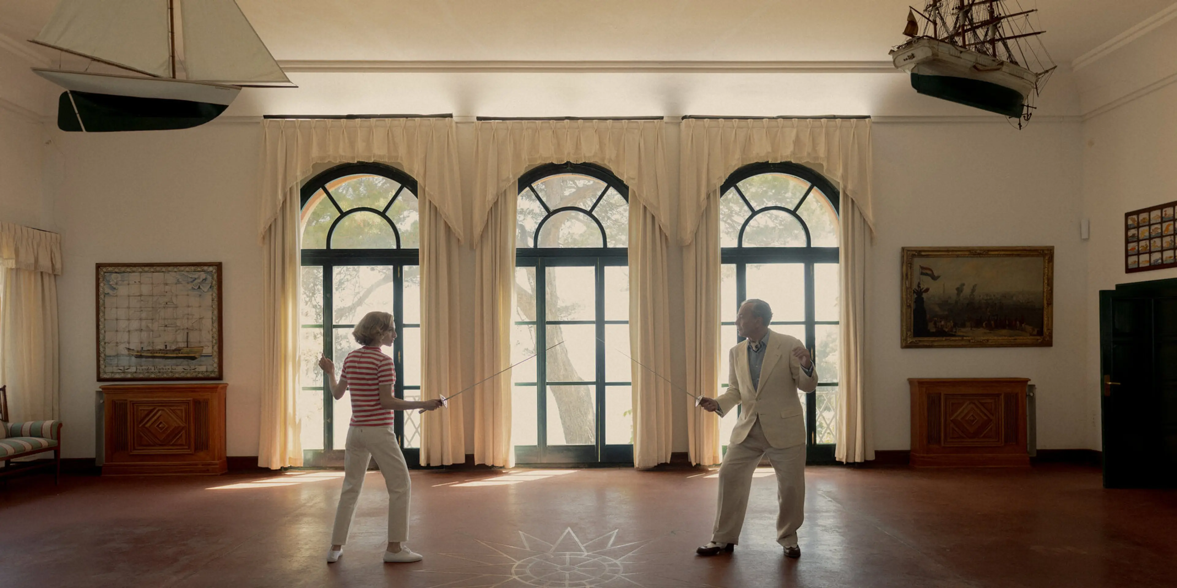 Basil and a woman practice fencing in a bright room with tall windows.