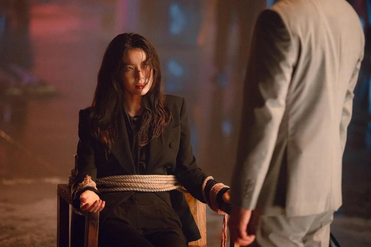Ha Young sits tied to a chair with injuries on her face, looking towards a man in a grey suit.