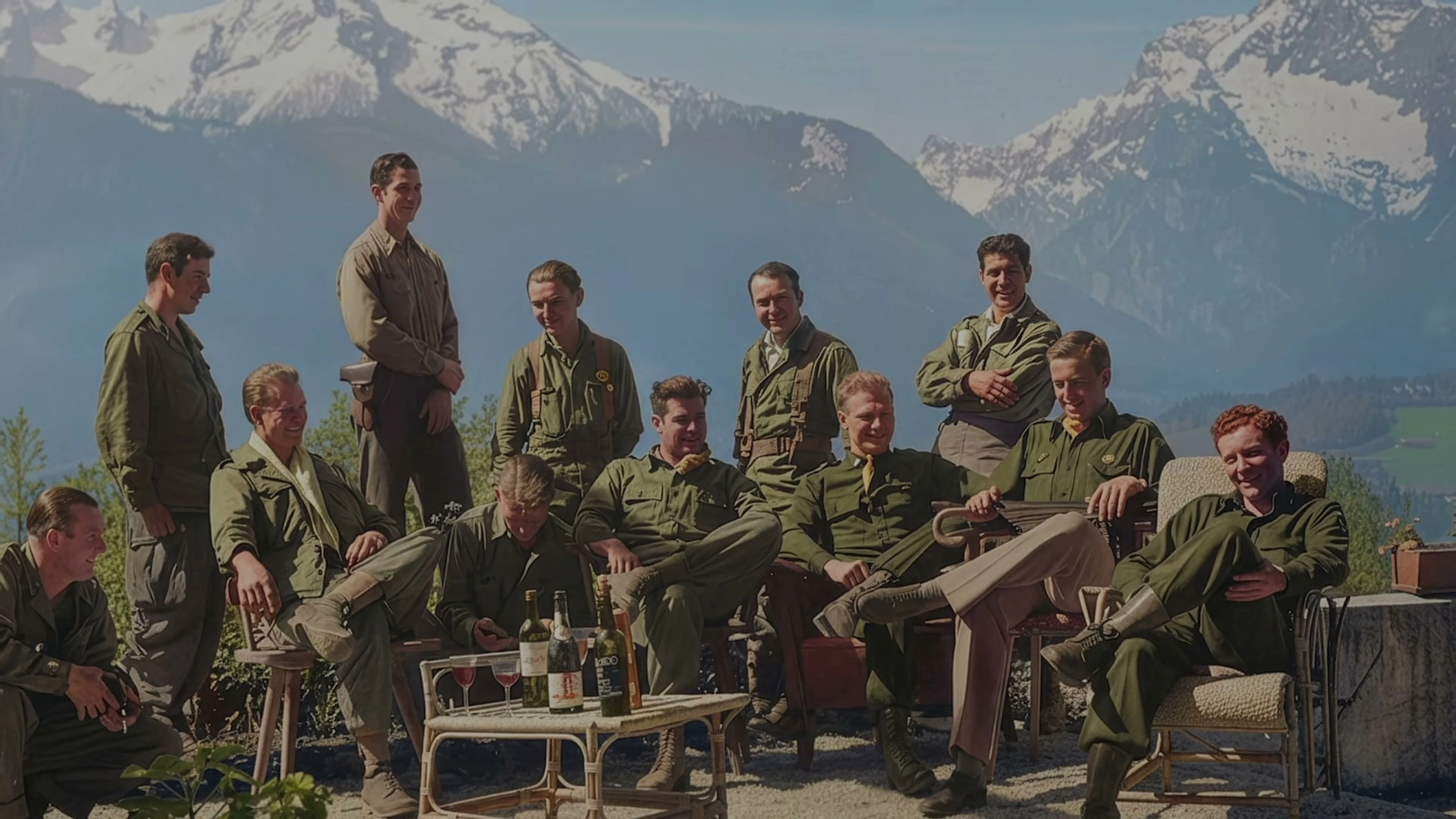 Members of the Band of Brothers cast gather outdoors in uniform with mountains behind them.