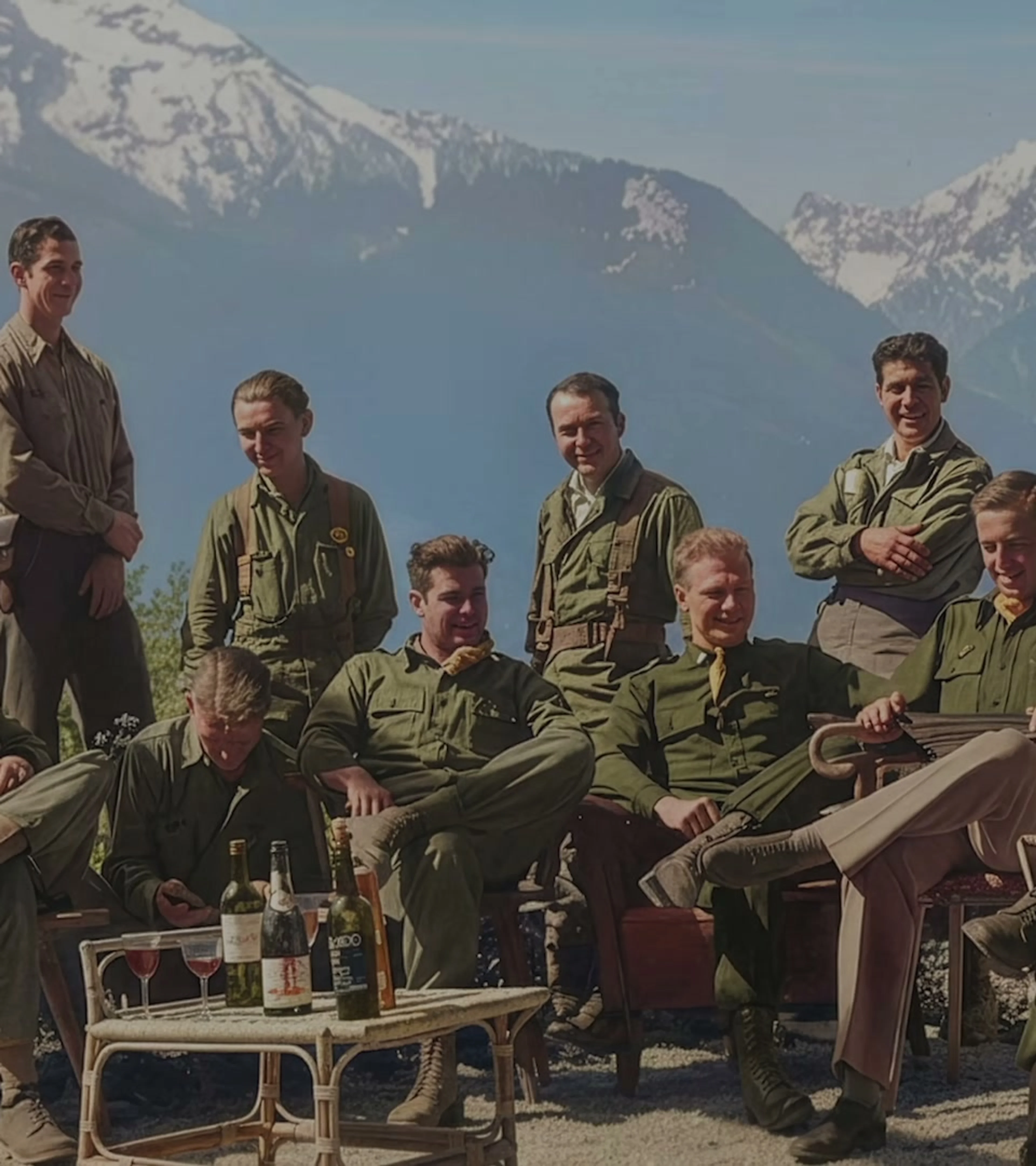 Members of the Band of Brothers cast gather outdoors in uniform with mountains behind them.