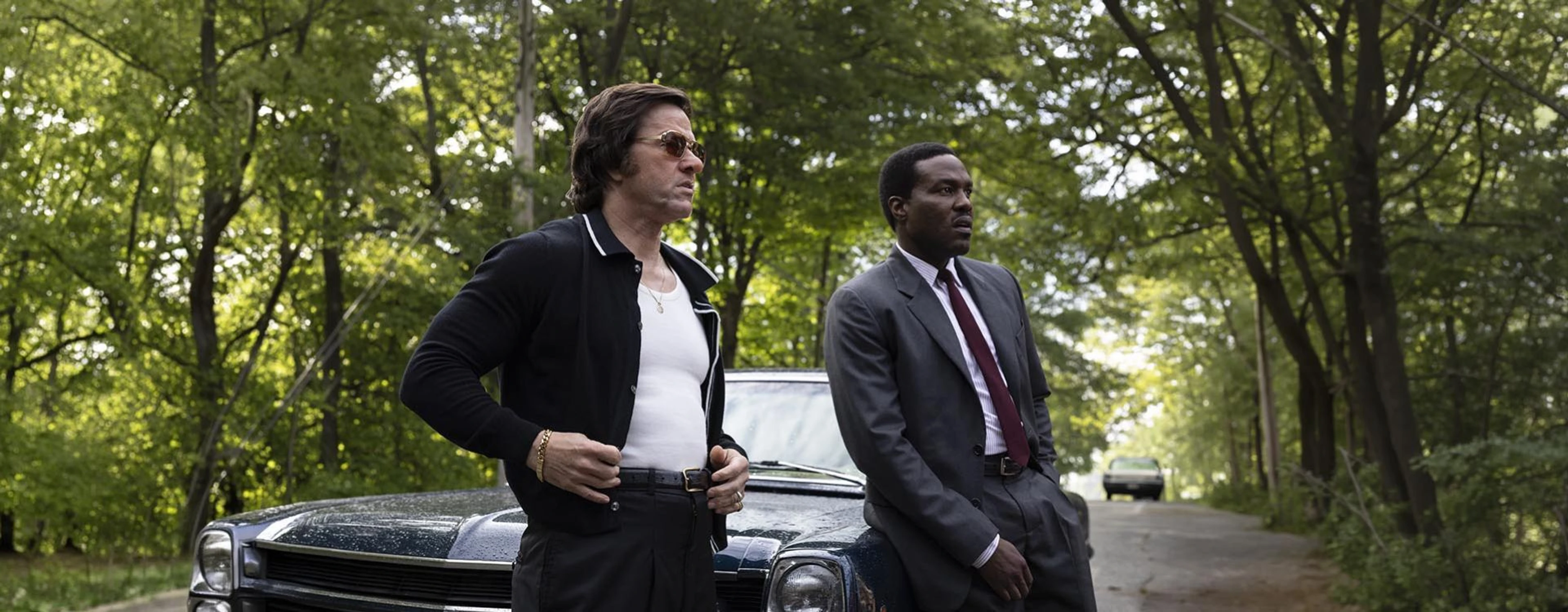 Yahya Abdul-Mateen II and Mark Wahlberg stand beside a car on a wooded road in By Any Means.