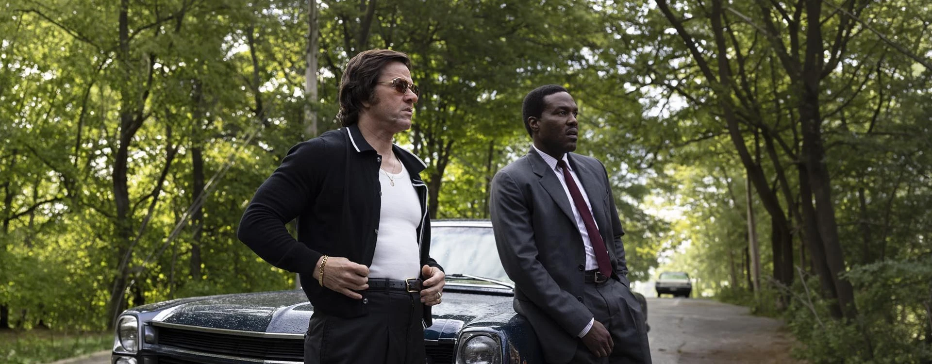 Yahya Abdul-Mateen II and Mark Wahlberg stand beside a car on a wooded road in By Any Means.