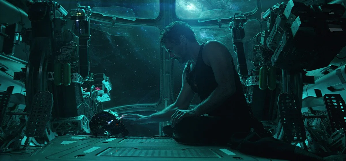 Robert Downey Jr. as Tony Stark sits beside his damaged Iron Man helmet aboard a dark spacecraft.
