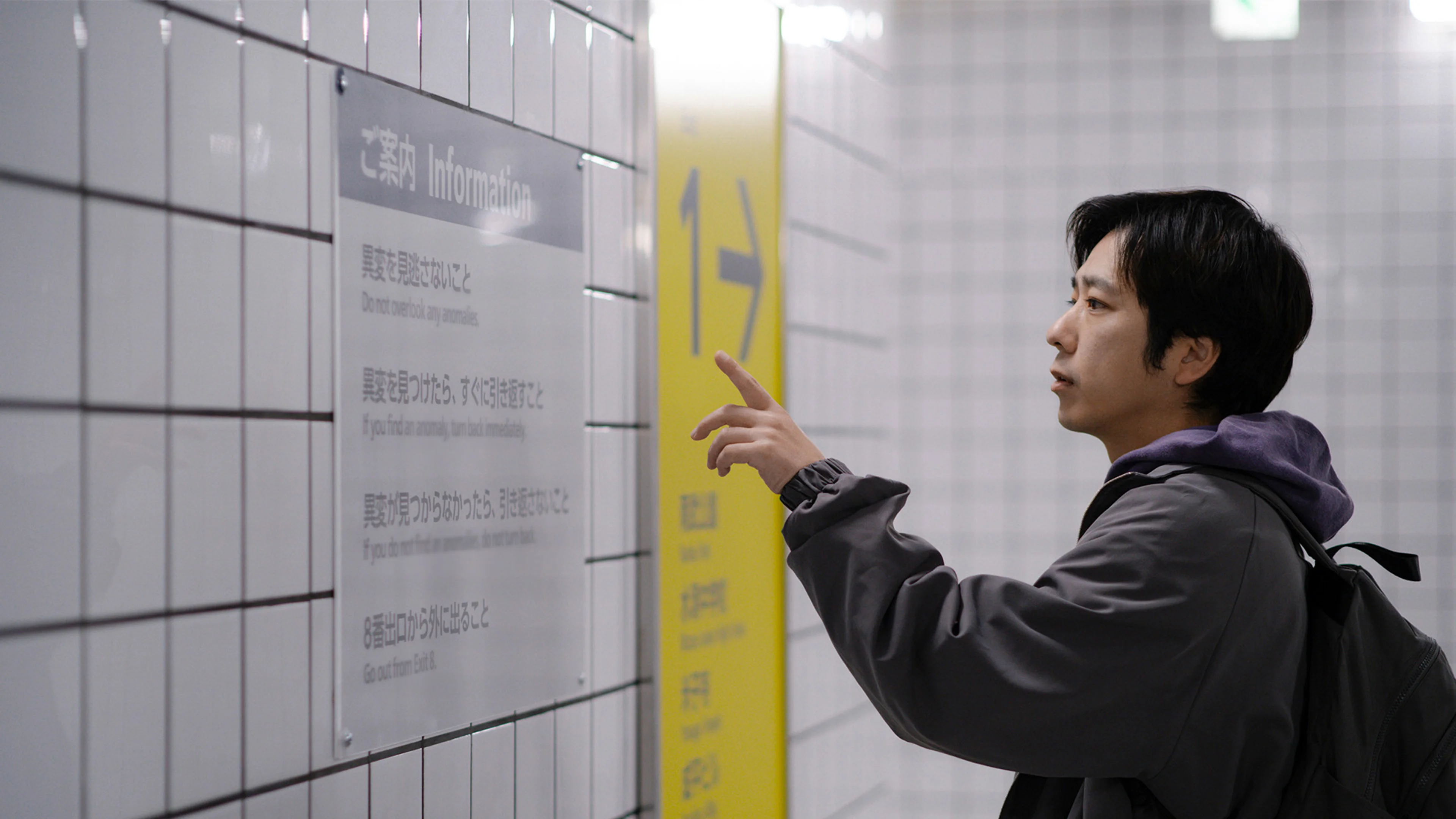 A man with a backpack points at the instructions displayed on a tiled wall.