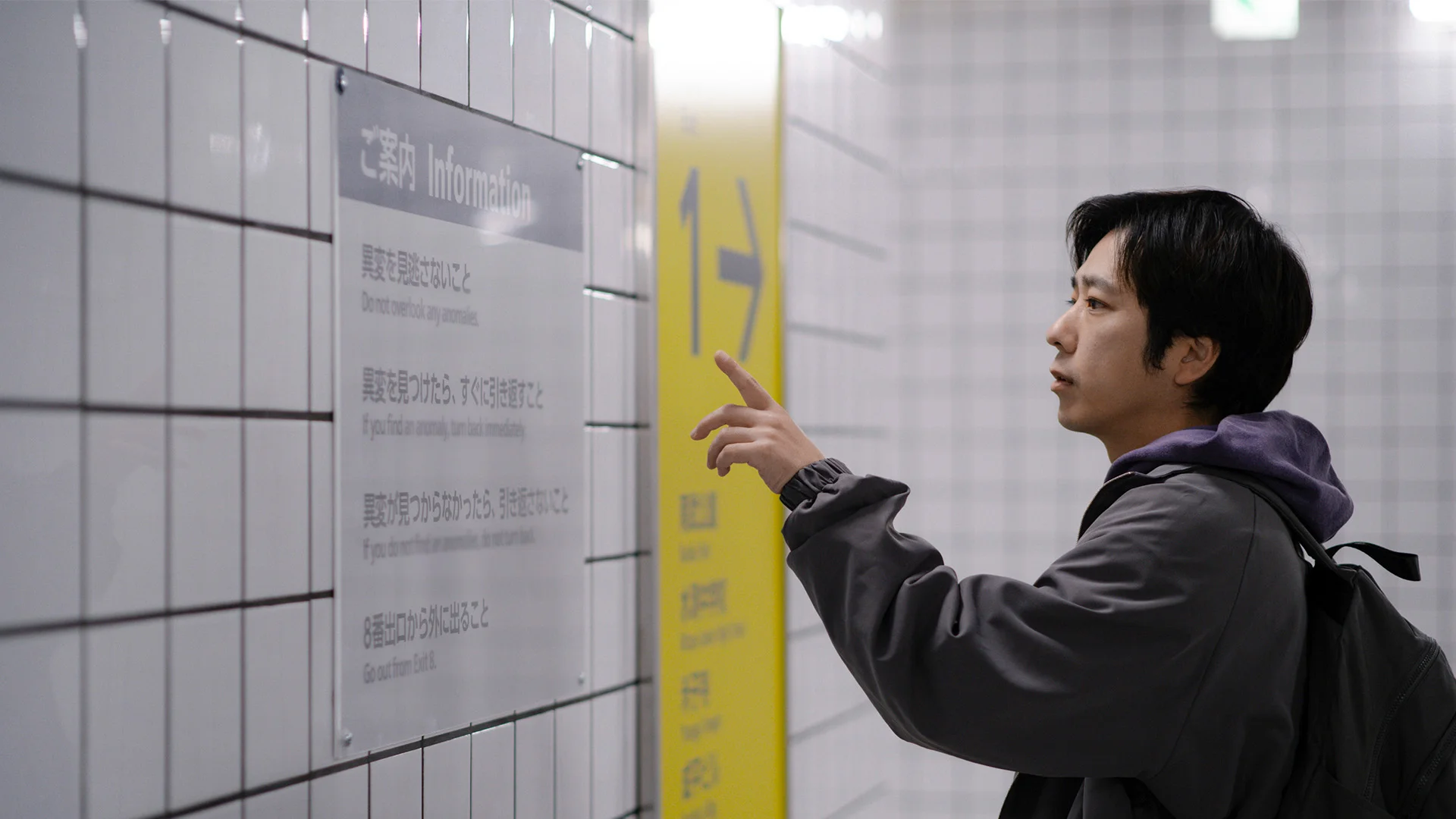 A man with a backpack points at the instructions displayed on a tiled wall.