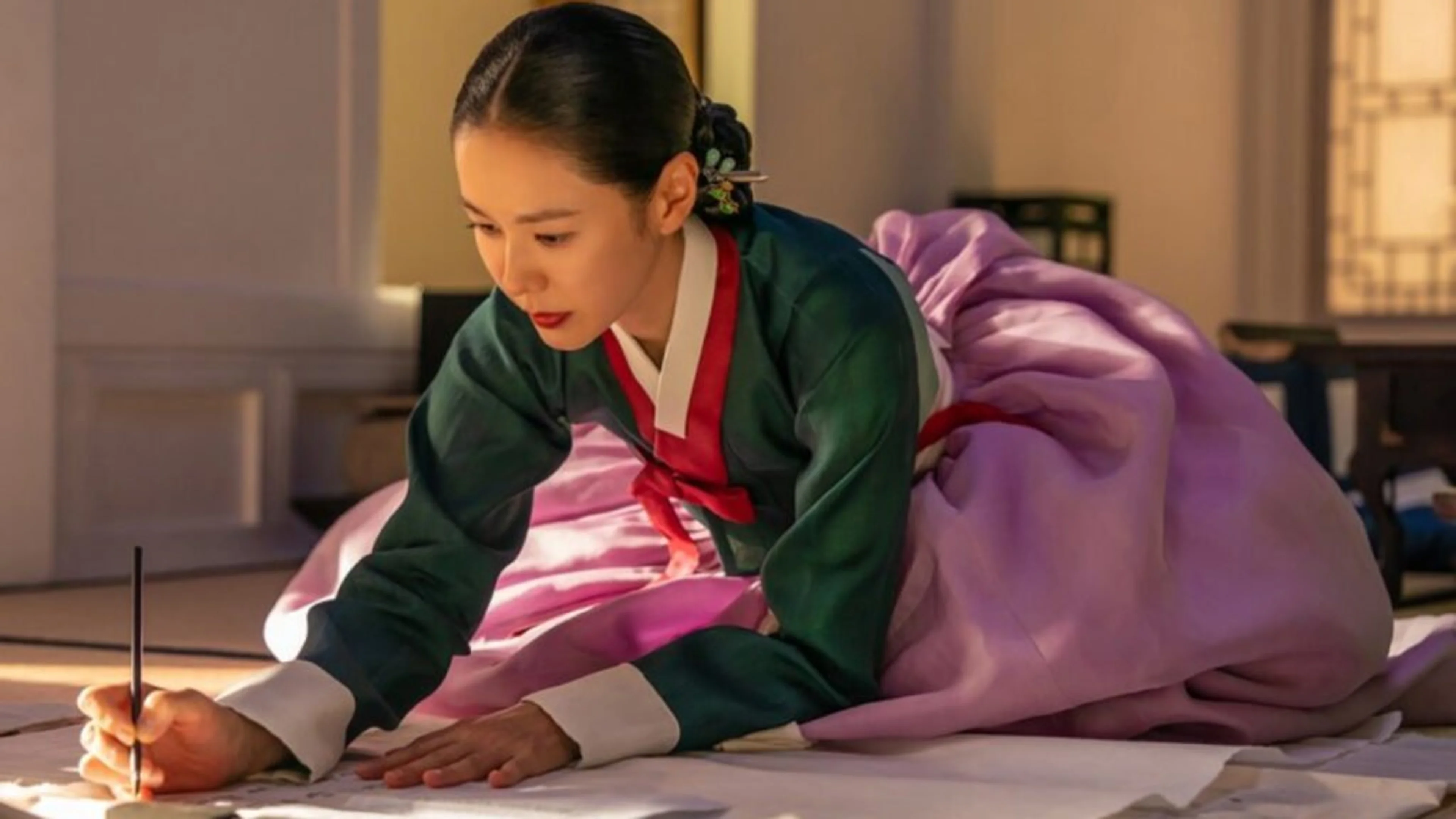 Lady Cho leans over paper with a writing brush in a sunlit room.