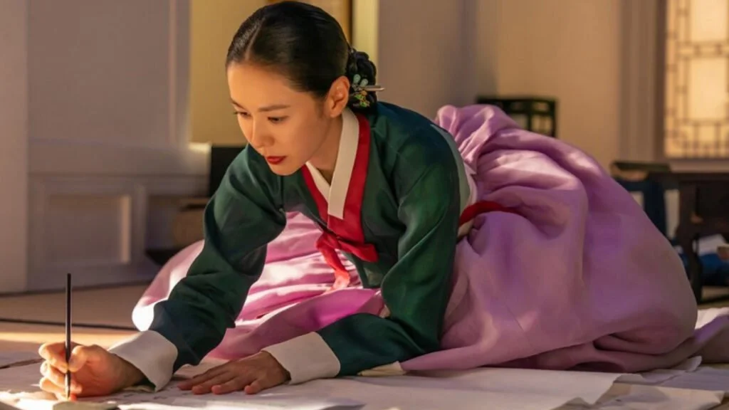Lady Cho leans over paper with a writing brush in a sunlit room.
