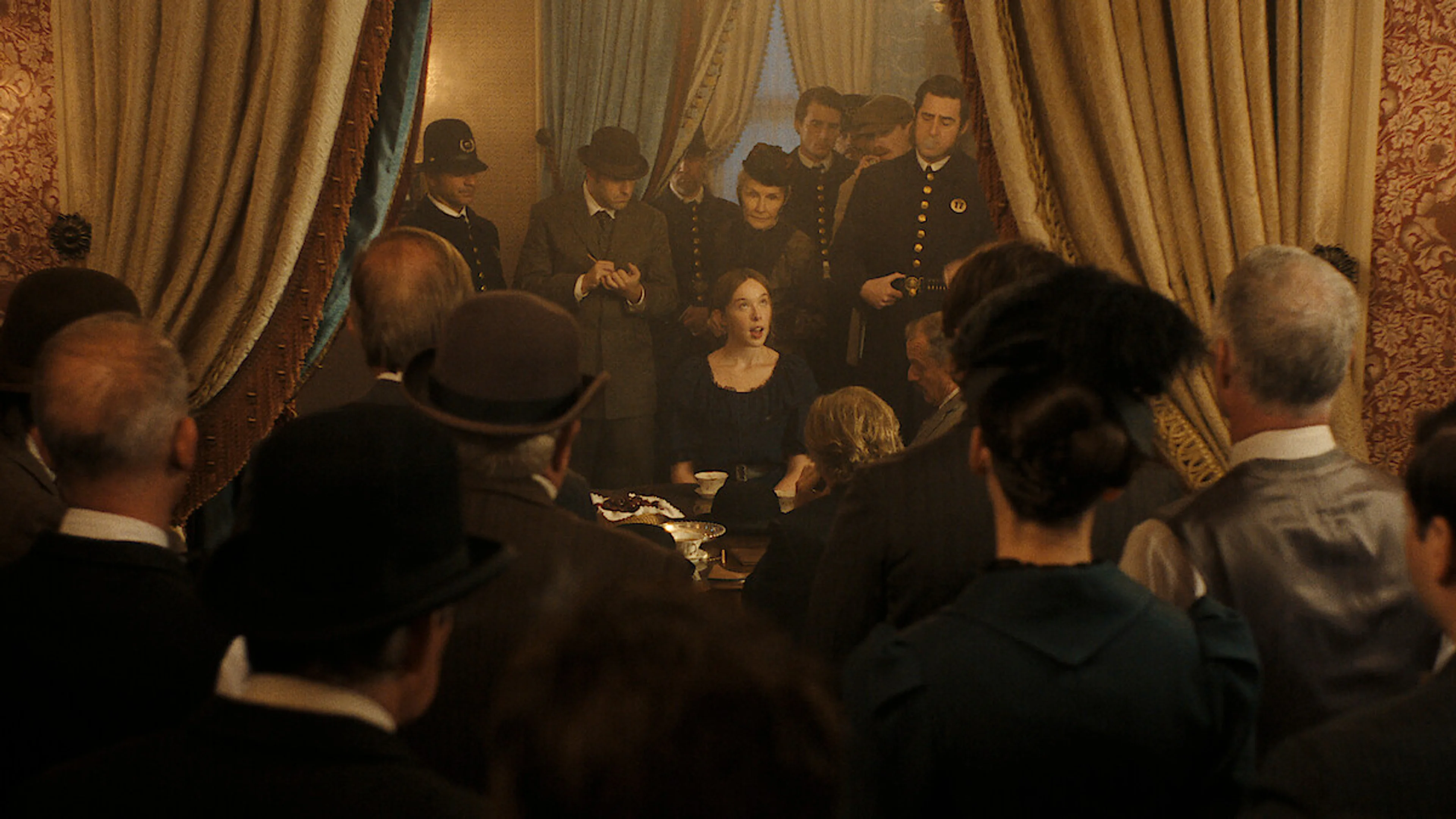 Lizzie sits at a crowded table surrounded by officers and onlookers in a period room.