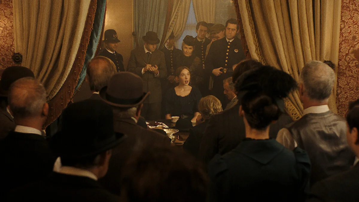 Lizzie sits at a crowded table surrounded by officers and onlookers in a period room.