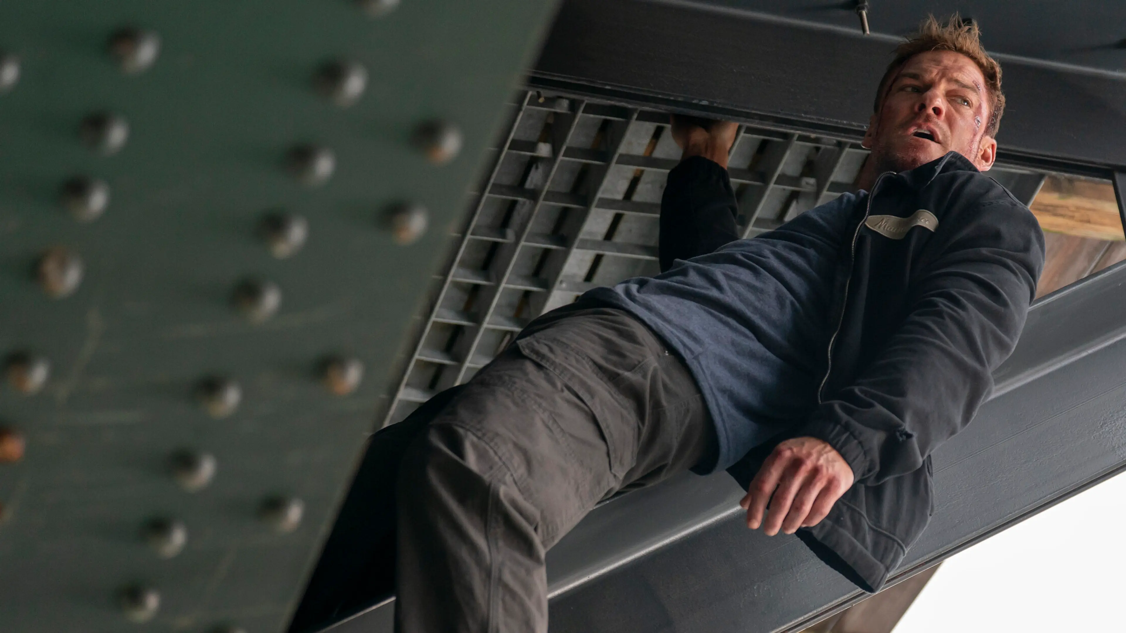 Alan Ritchson as Jack Reacher hanging from a steel structure during an action scene