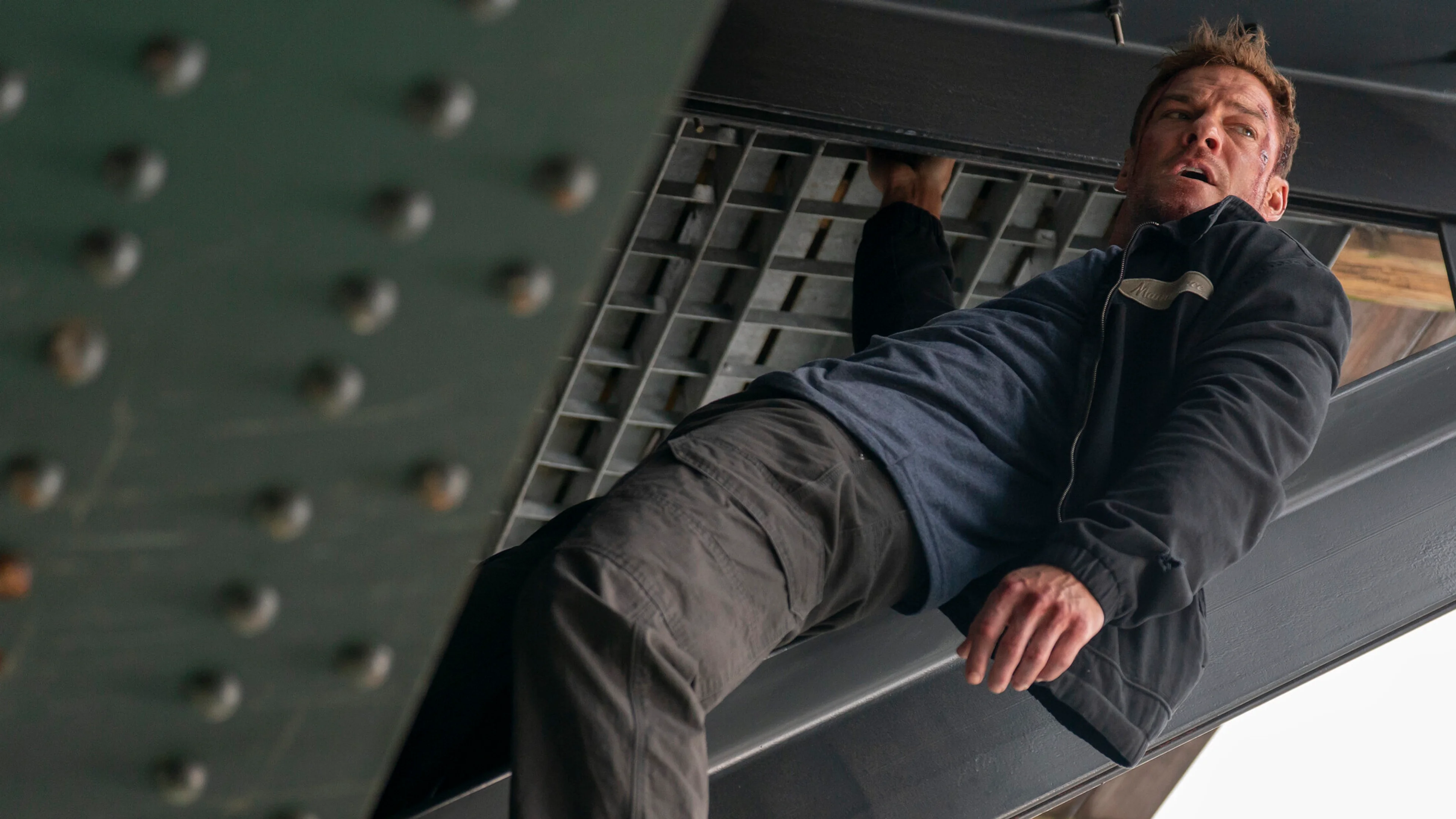 Alan Ritchson as Jack Reacher hanging from a steel structure during an action scene