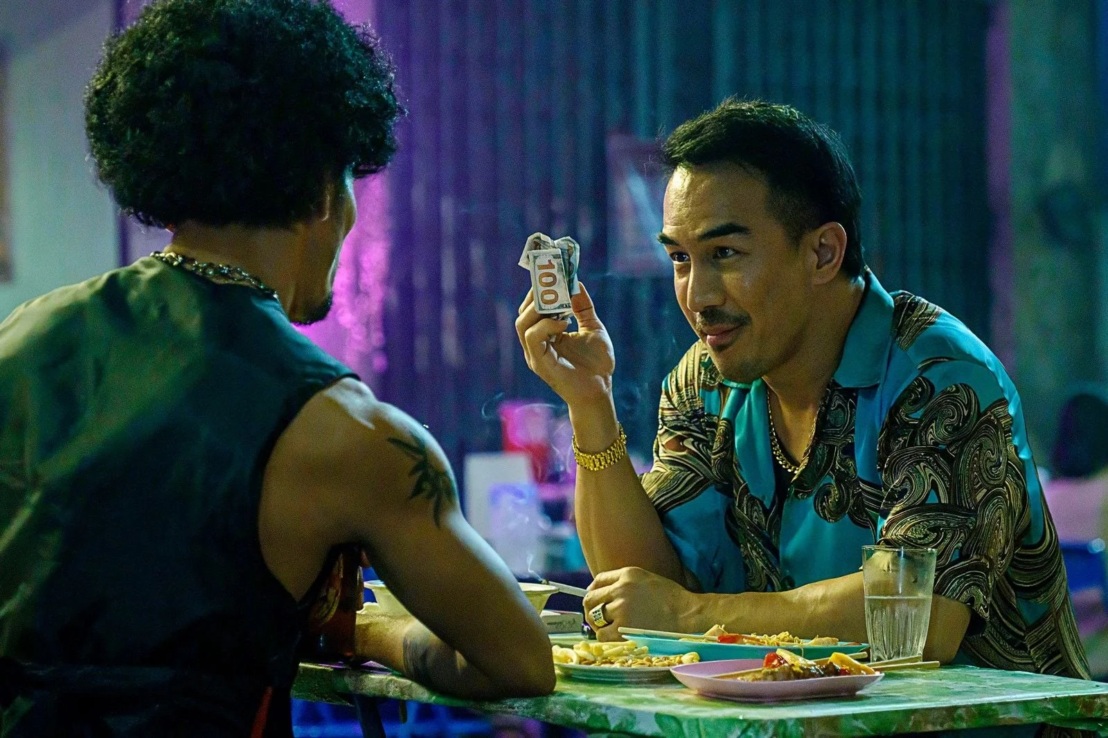 Joe Taslim as Navin holds a banknote during a conversation at a table.