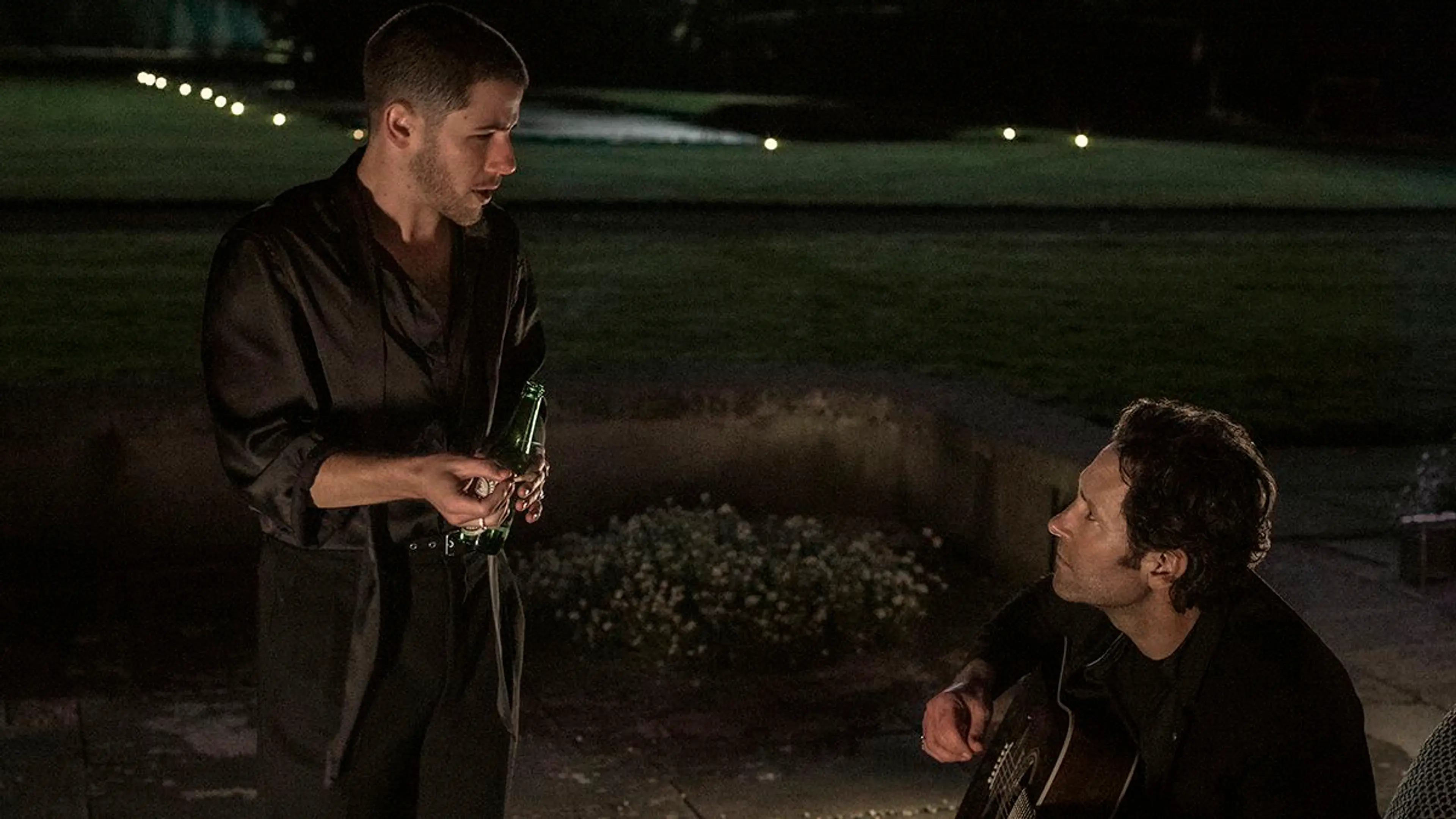 Danny stands holding a bottle while Rick sits playing an acoustic guitar on a patio at night.