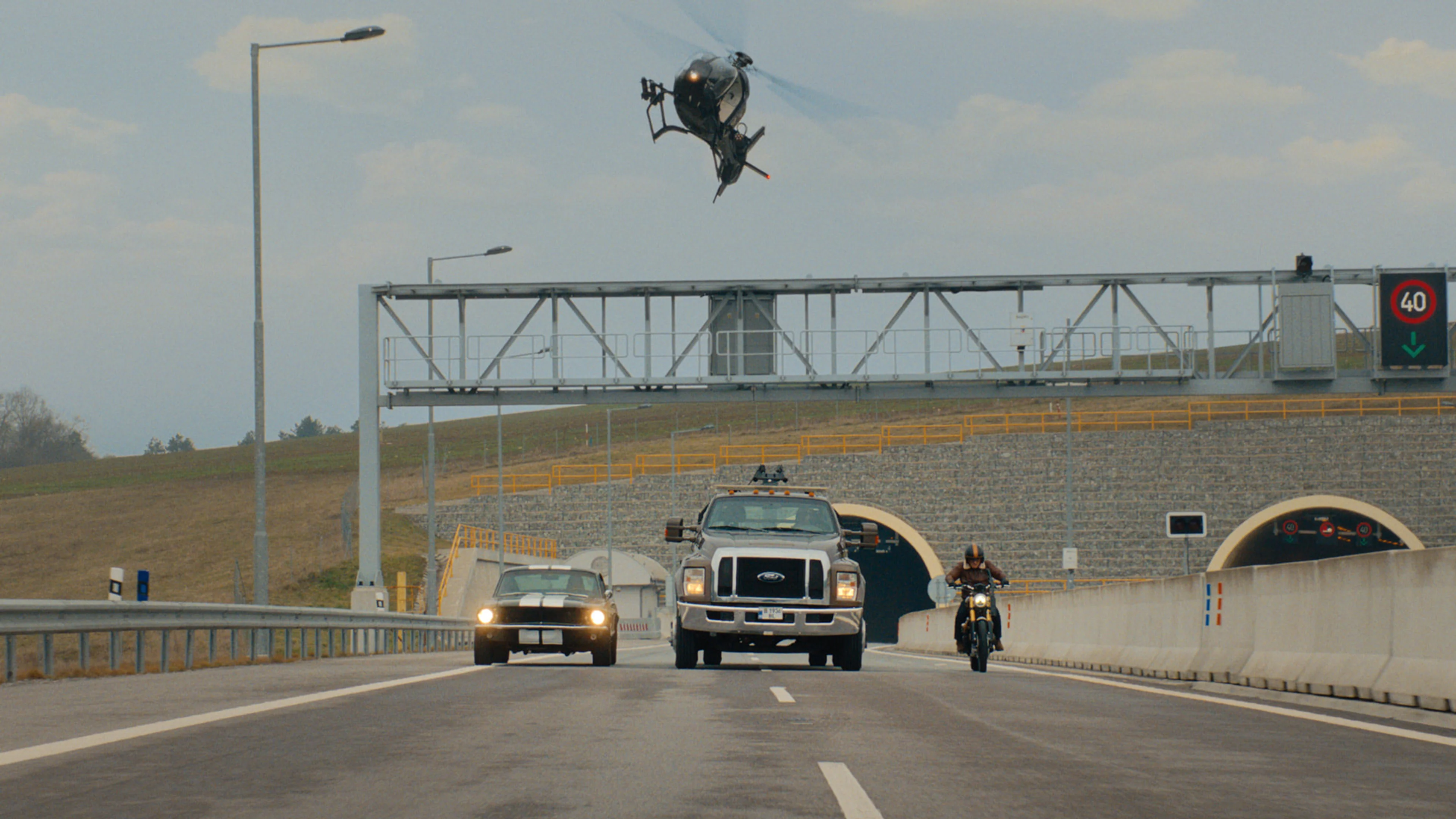 Cars and a motorcycle race along a road beneath a helicopter in Matchbox The Movie.