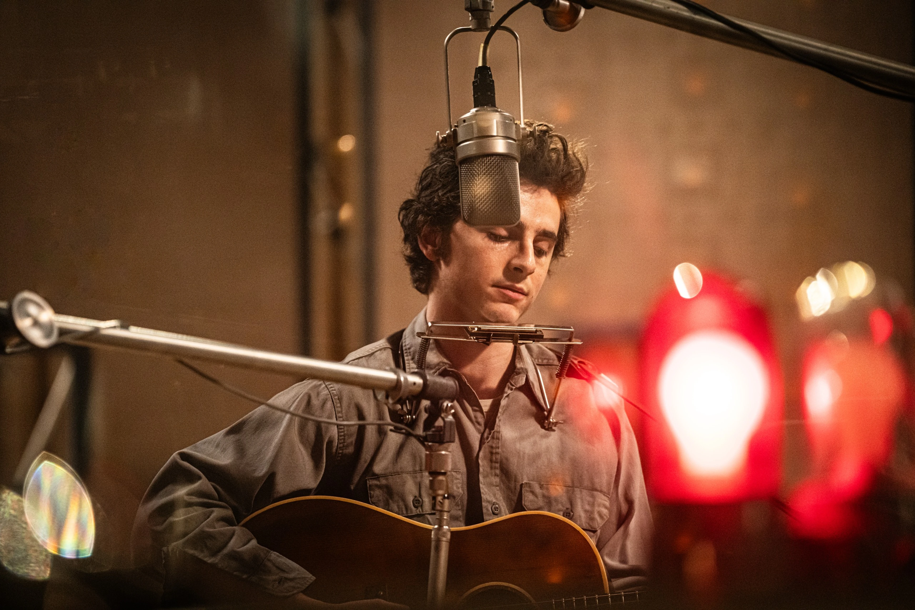 Timothée Chalamet as Bob Dylan plays acoustic guitar in front of a studio microphone.