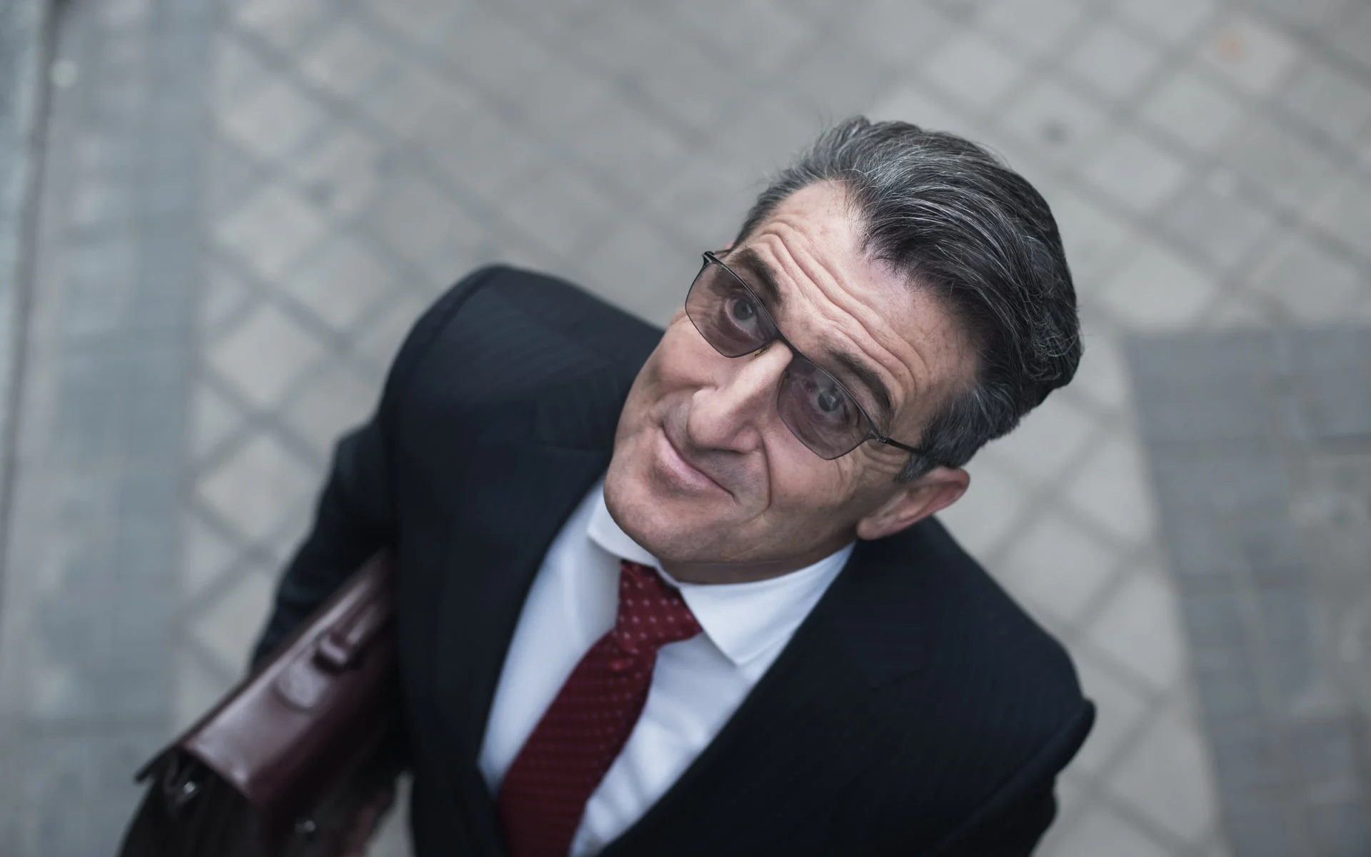 A suited man wearing glasses leans toward someone out of frame in an official Collision Course still.