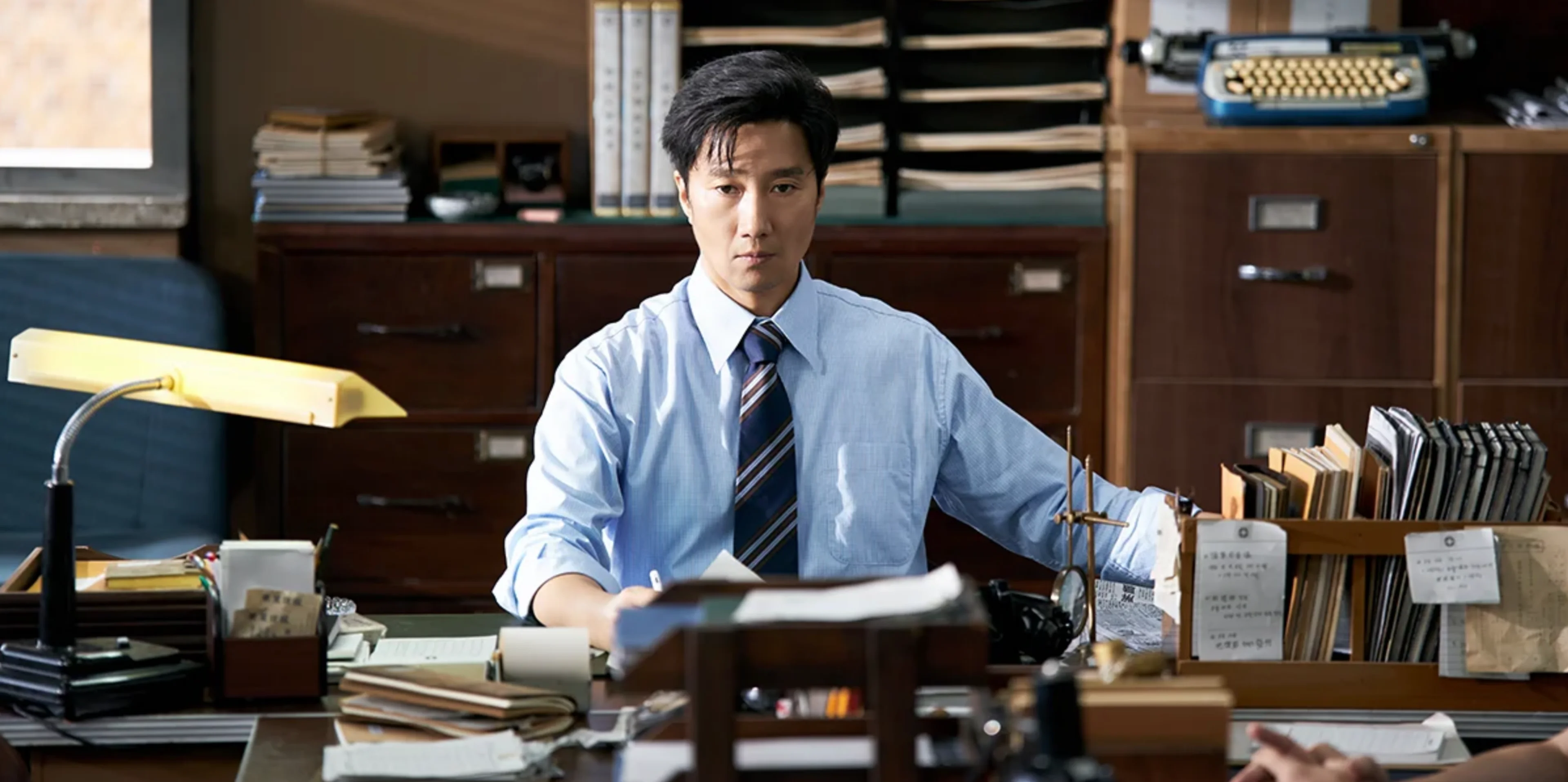 Park Hae-il as editor Jae-hwan sits behind a desk covered with papers and folders.
