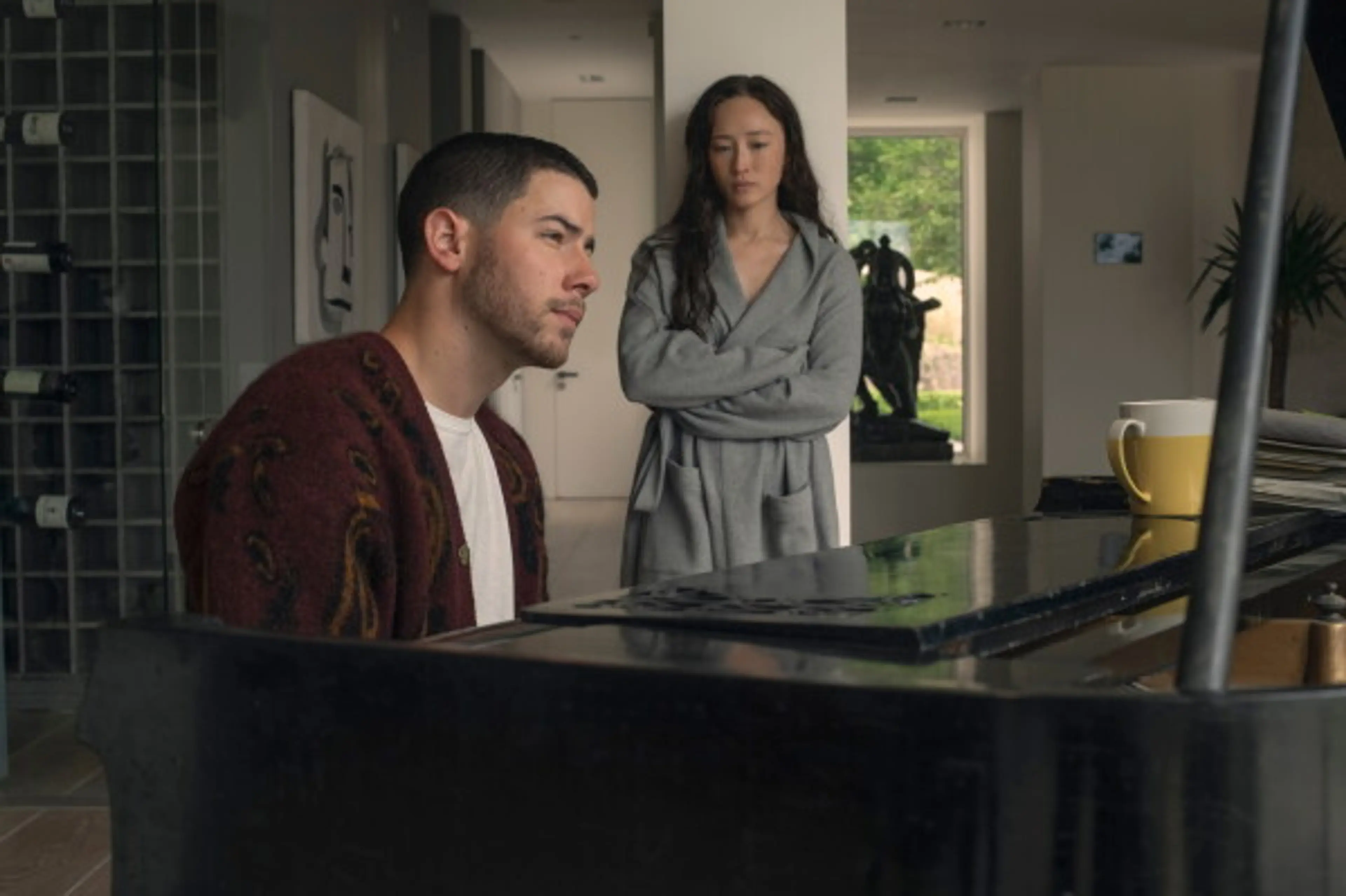 Danny sits at a piano while Marcia stands behind him with her arms crossed.