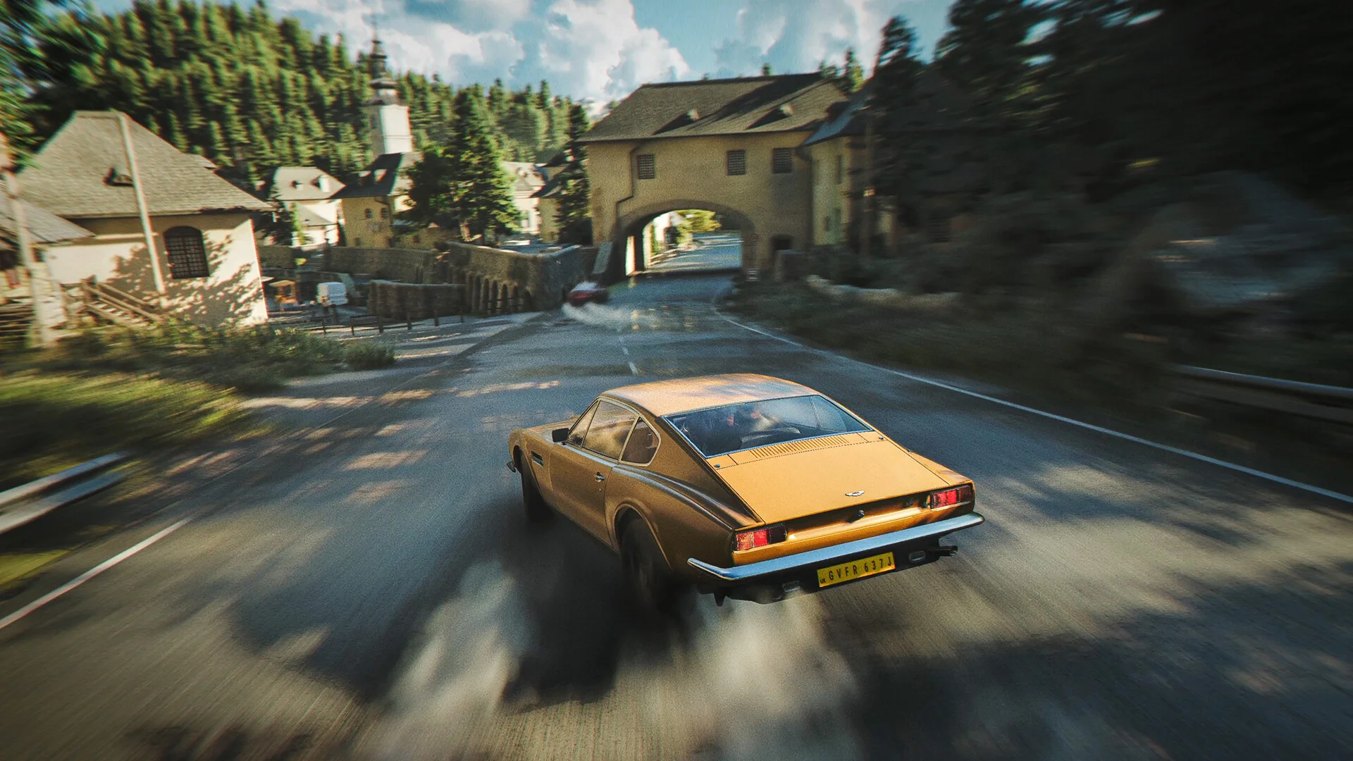 A gold Aston Martin drives along a village road beneath mountains.
