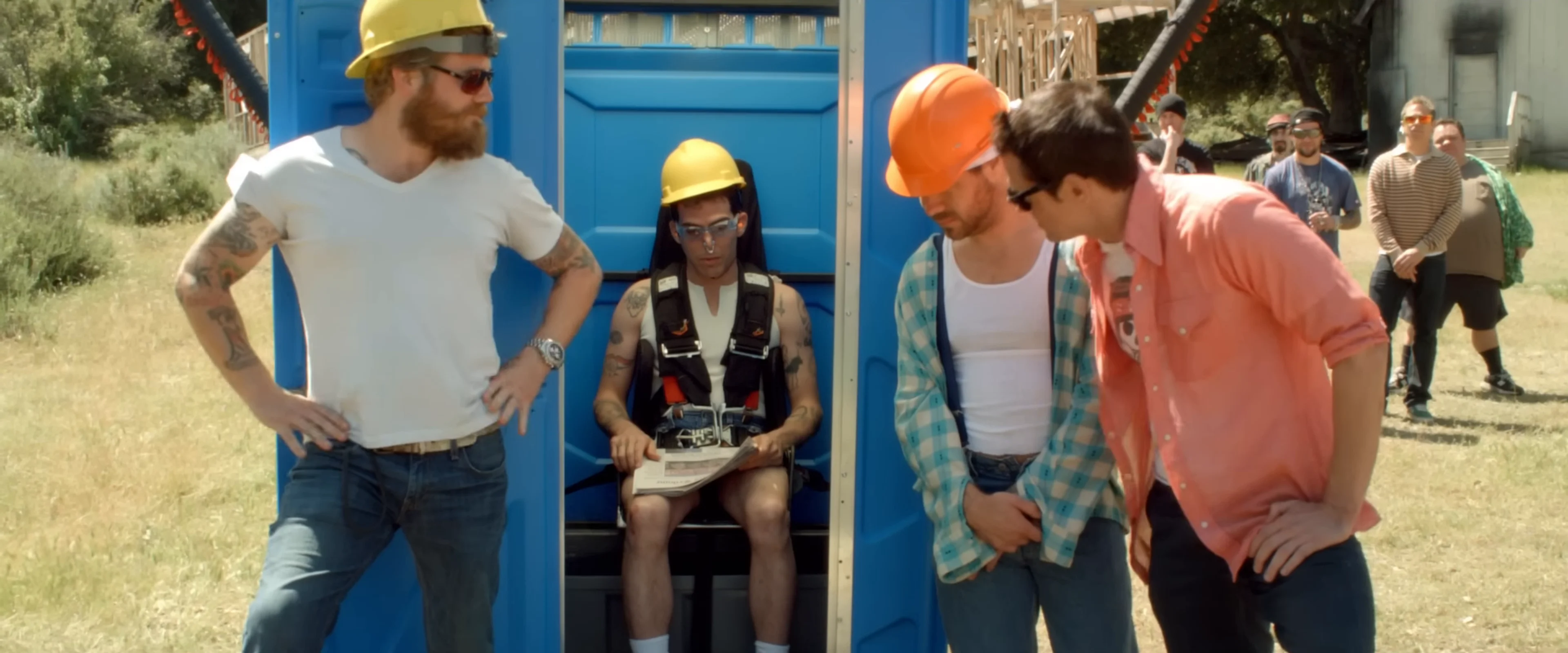 Several Jackass cast members gather around a blue booth on an outdoor stunt set.