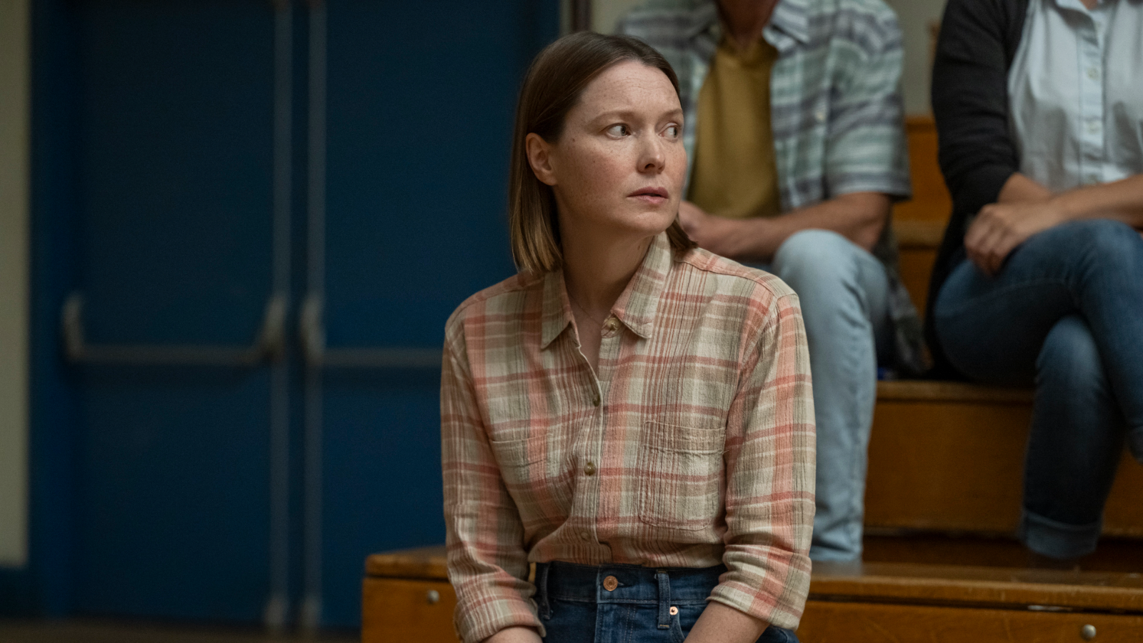 Samantha Sloyan as Margaret White sits against blue bleachers and looks to her left.