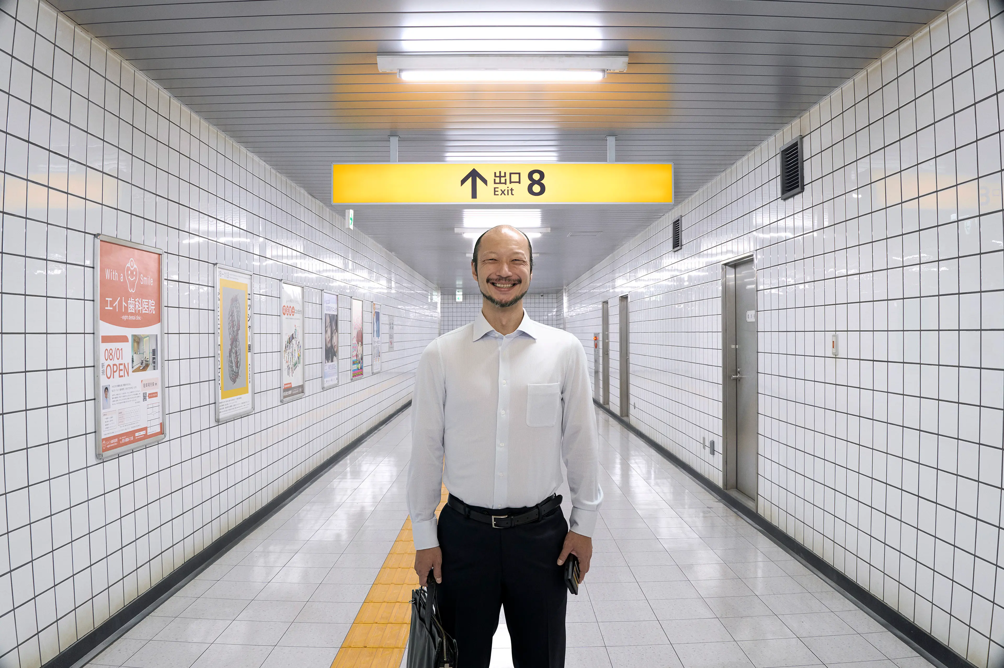 A man in a white shirt smiles in a tiled subway corridor beneath a yellow Exit 8 sign.