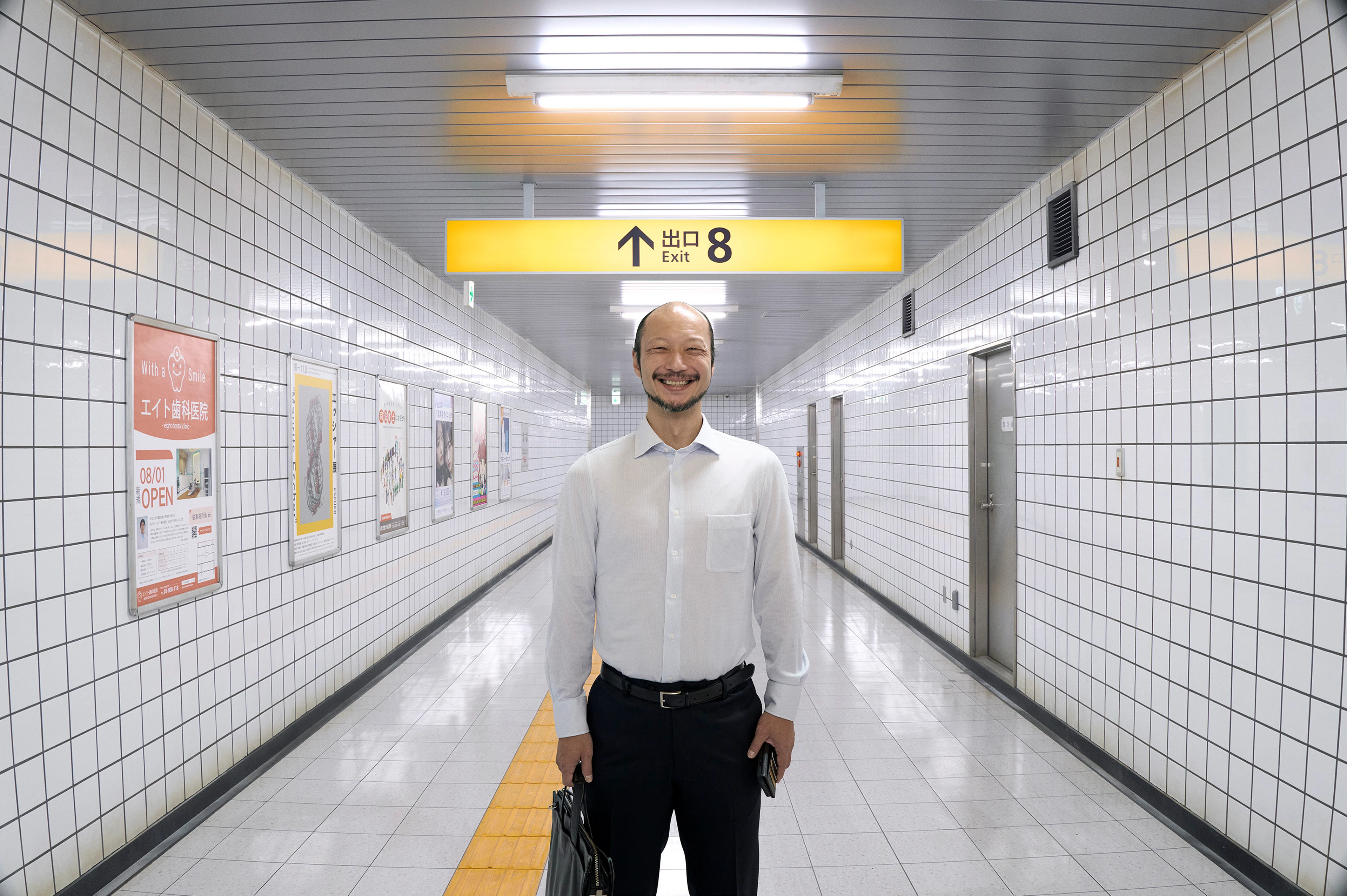 A man in a white shirt smiles in a tiled subway corridor beneath a yellow Exit 8 sign.