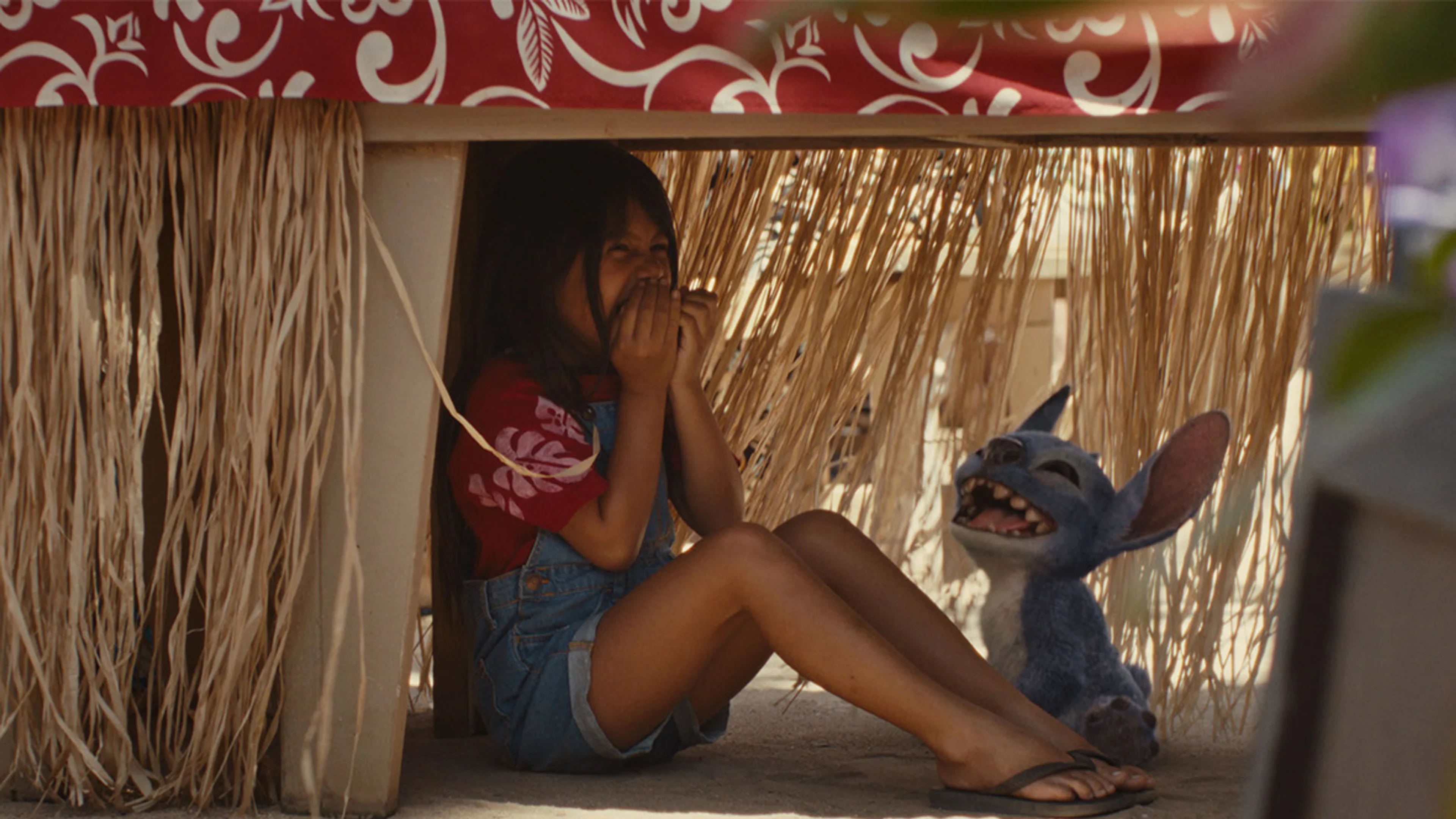 Lilo and Stitch laugh together while hiding under a table in the 2025 live-action Lilo & Stitch film.