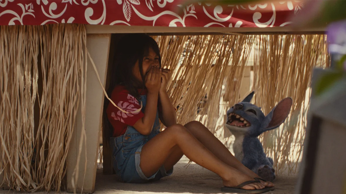 Lilo and Stitch laugh together while hiding under a table in the 2025 live-action Lilo & Stitch film.